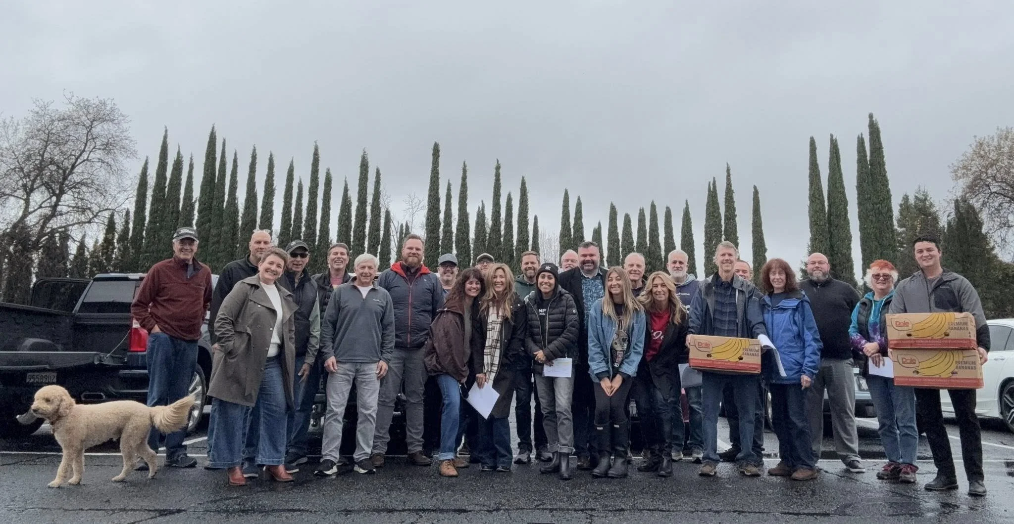 Group of people standing outdoors on a cloudy day, some holding boxes of bananas, with a dog, cars, and tall trees in the background.