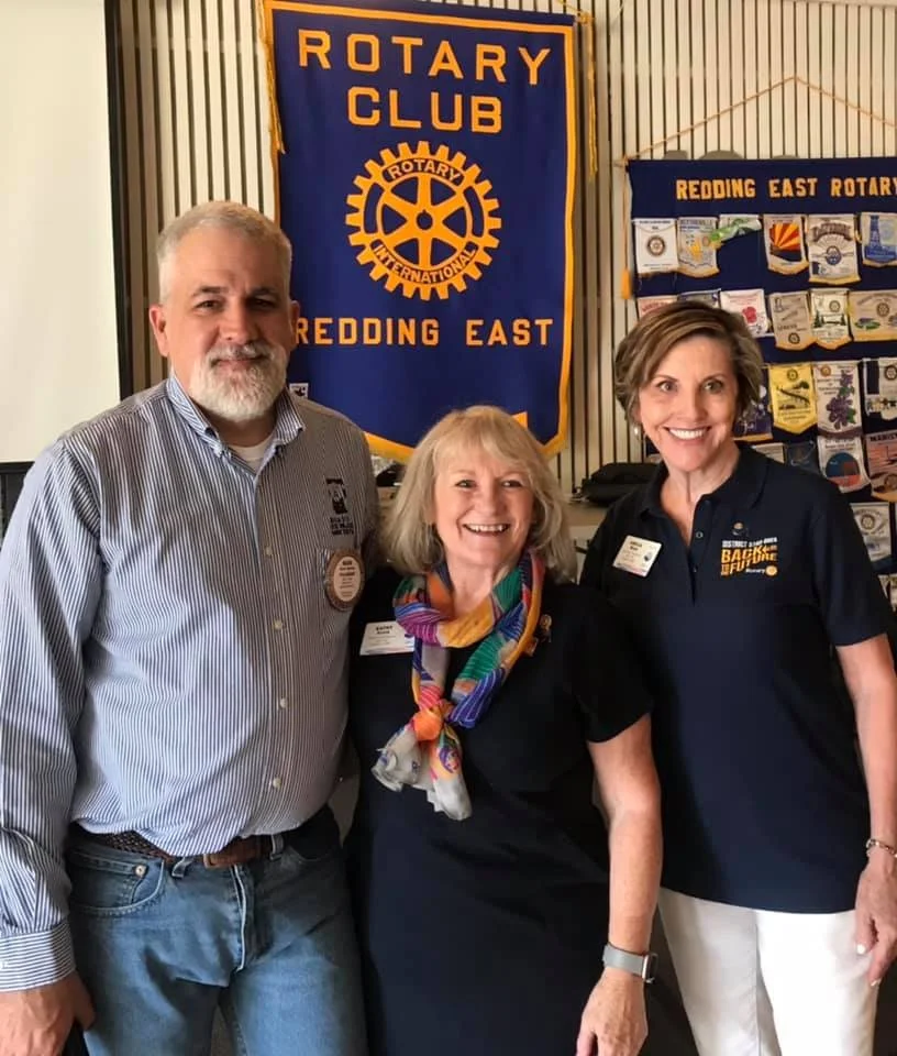 Three people standing together in front of Rotary Club banners, smiling for the photo. One man on the left wearing a striped shirt, two women on the right wearing dark shirts, one with a colorful scarf, in an indoor setting with display boards in the