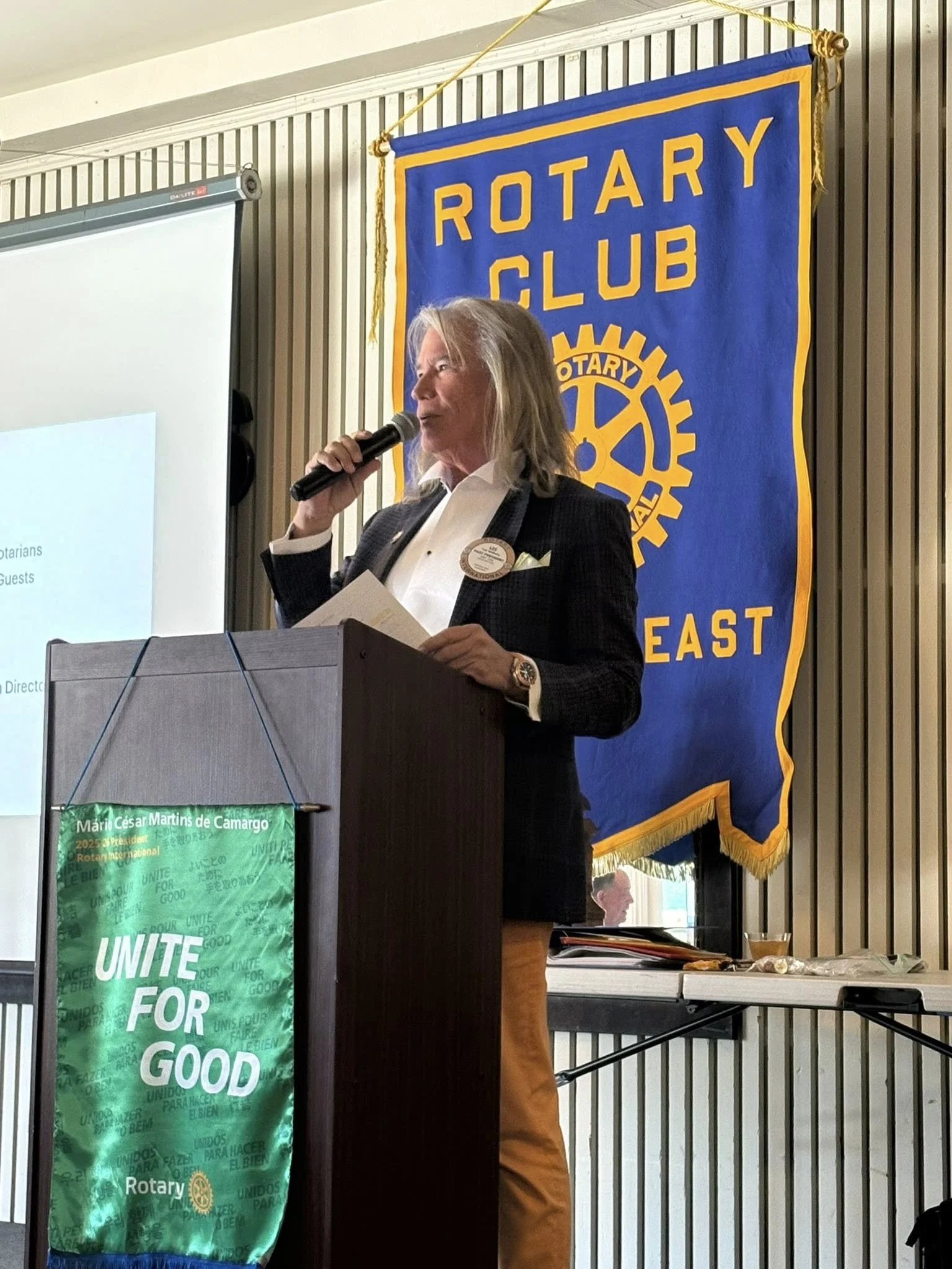 A woman standing at a podium with a Rotary Club banner behind her, speaking into a microphone at a formal meeting.