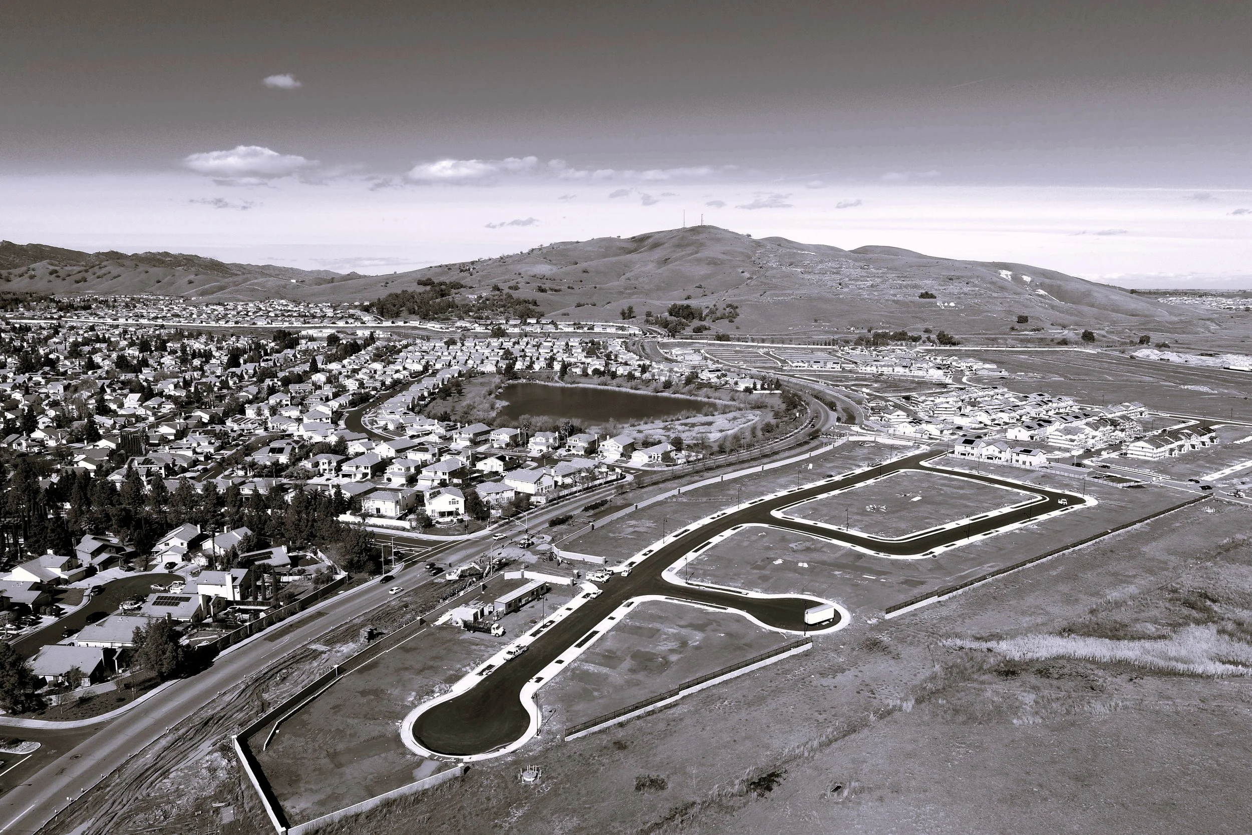 A black and white aerial view of a residential neighborhood with houses, streets, a small pond, and surrounding hills in the distance.