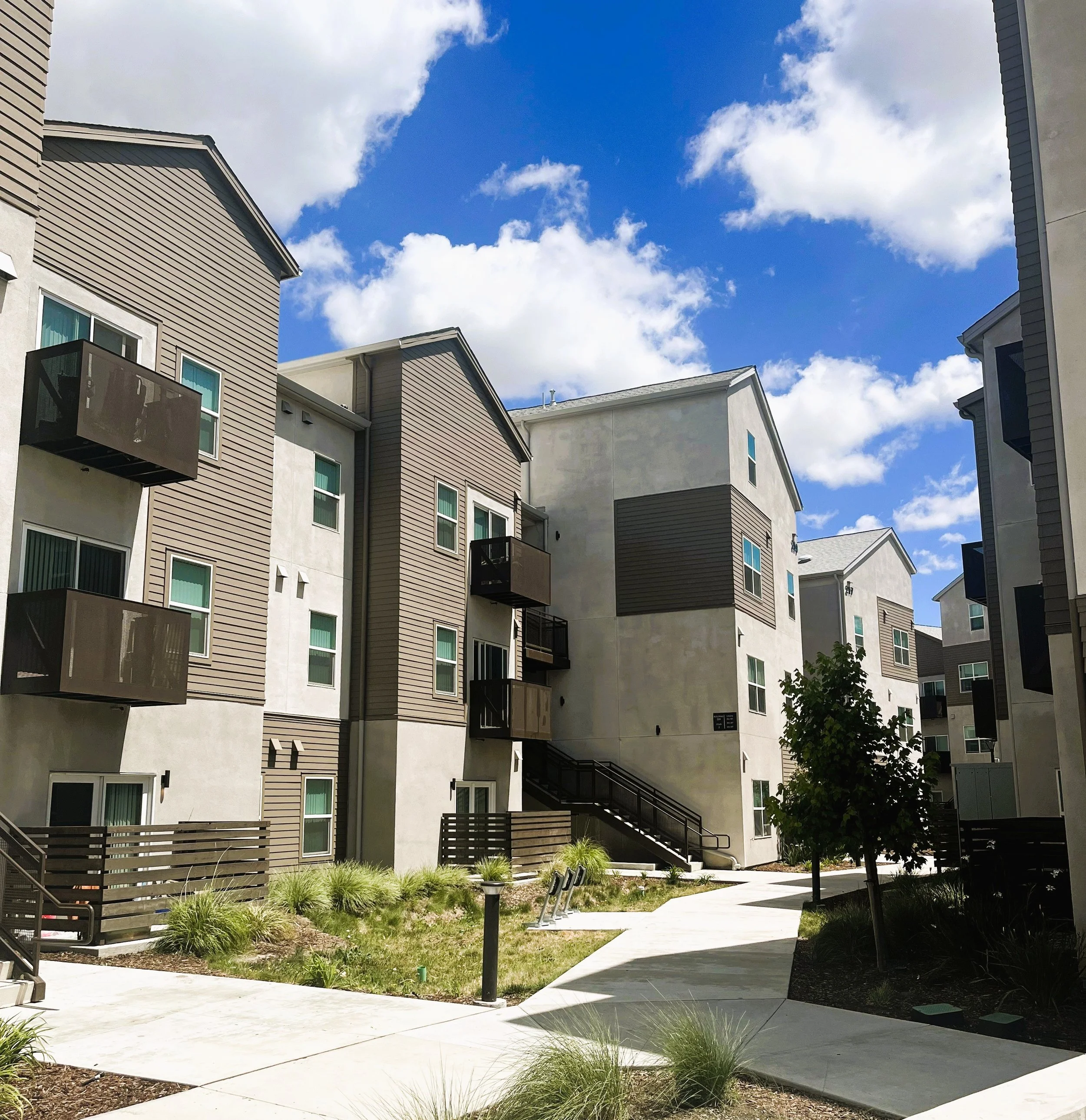 Modern residential apartment buildings with multiple stories, beige and gray siding, balconies, and a courtyard with a sidewalk, small plants, and a tree, under a partly cloudy sky.