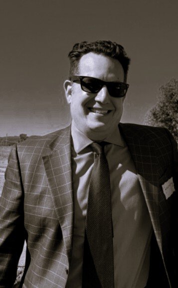 A smiling man in a checkered suit and sunglasses outdoors, with a clear sky and trees in the background.