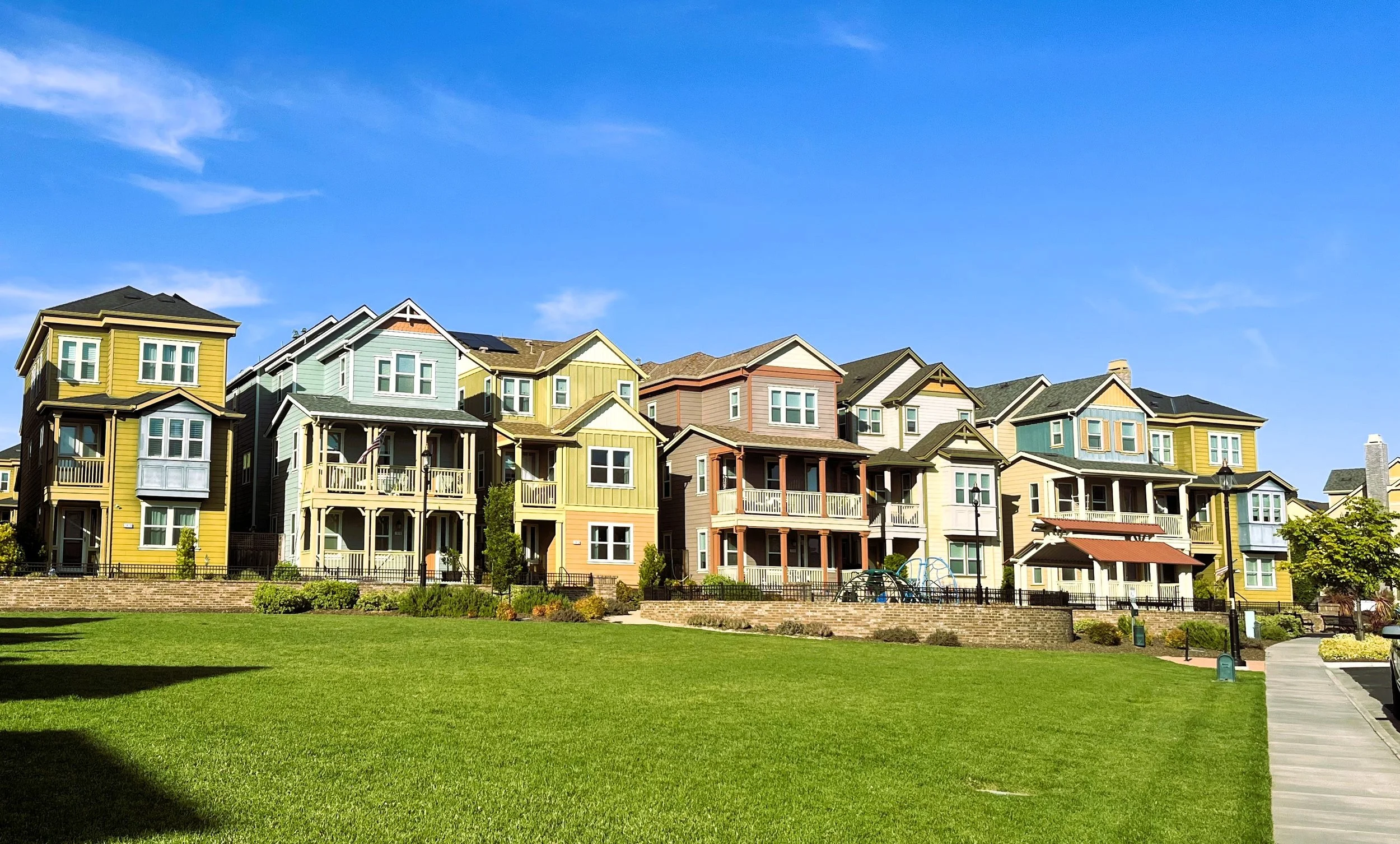A row of colorful multi-story houses with balconies, set against a bright blue sky, with a well-maintained green lawn and a walking path in the foreground.