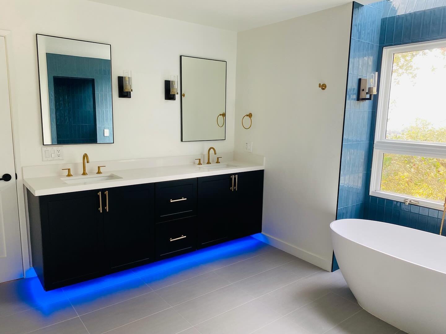 Modern bathroom with black vanity and two white sinks, gold fixtures, large mirrors, blue tiled section around window, freestanding bathtub, and blue LED lighting under vanity.