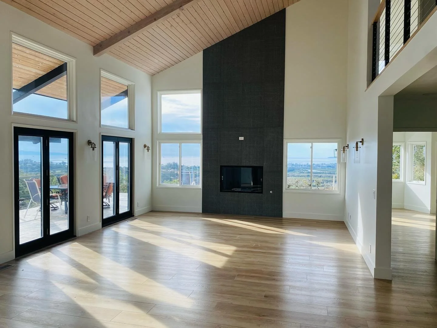 Bright living room with high vaulted wooden ceiling, large windows, and glass doors leading to a balcony. Modern fireplace on a dark accent wall, light hardwood flooring, with outdoor patio furniture visible outside.