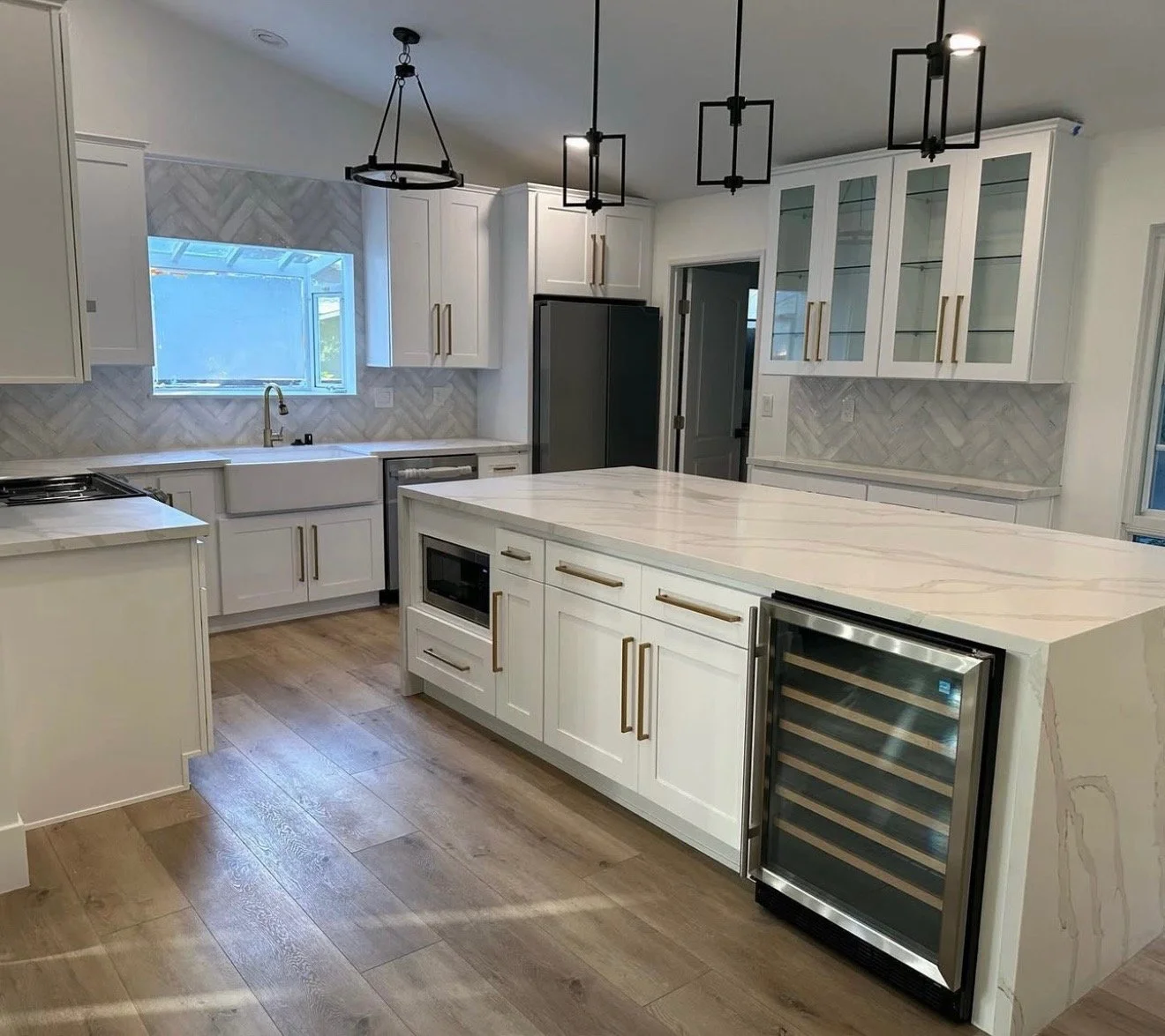 Modern kitchen with white cabinets, marble countertop island, wooden flooring, stainless steel appliances, window above the sink, glass-front cabinets, and three contemporary pendant lights hanging from the ceiling.
