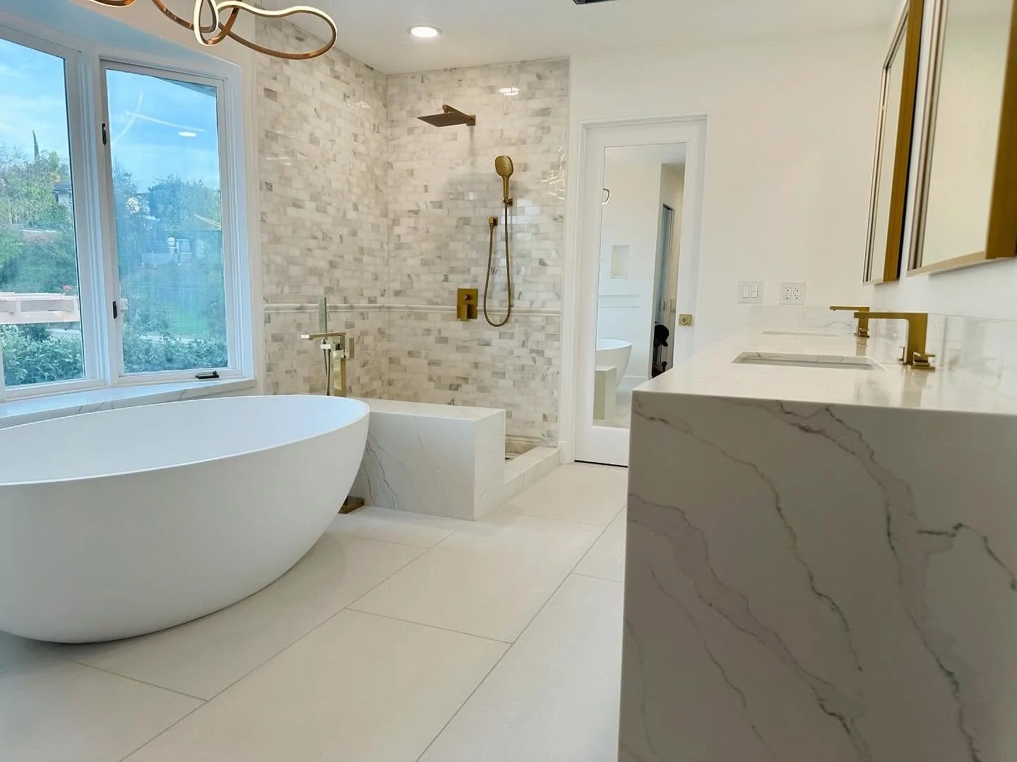 Modern bathroom with large freestanding bathtub, walk-in shower with gold fixtures, and double vanity with marble countertop and gold faucets. Large window lets in natural light.