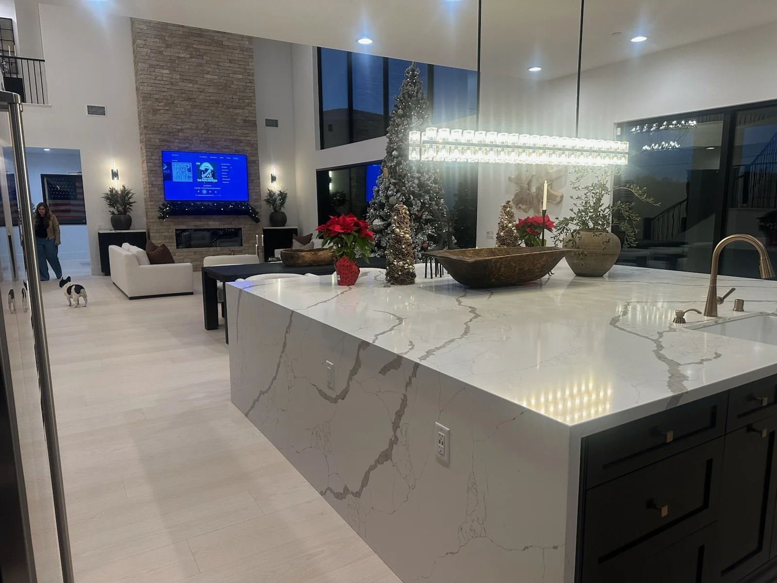 Modern open-concept living space with a kitchen island, decorated for Christmas, including a Christmas tree, poinsettias, and festive ornaments, with a living room featuring a fireplace, large TV, and seating area.