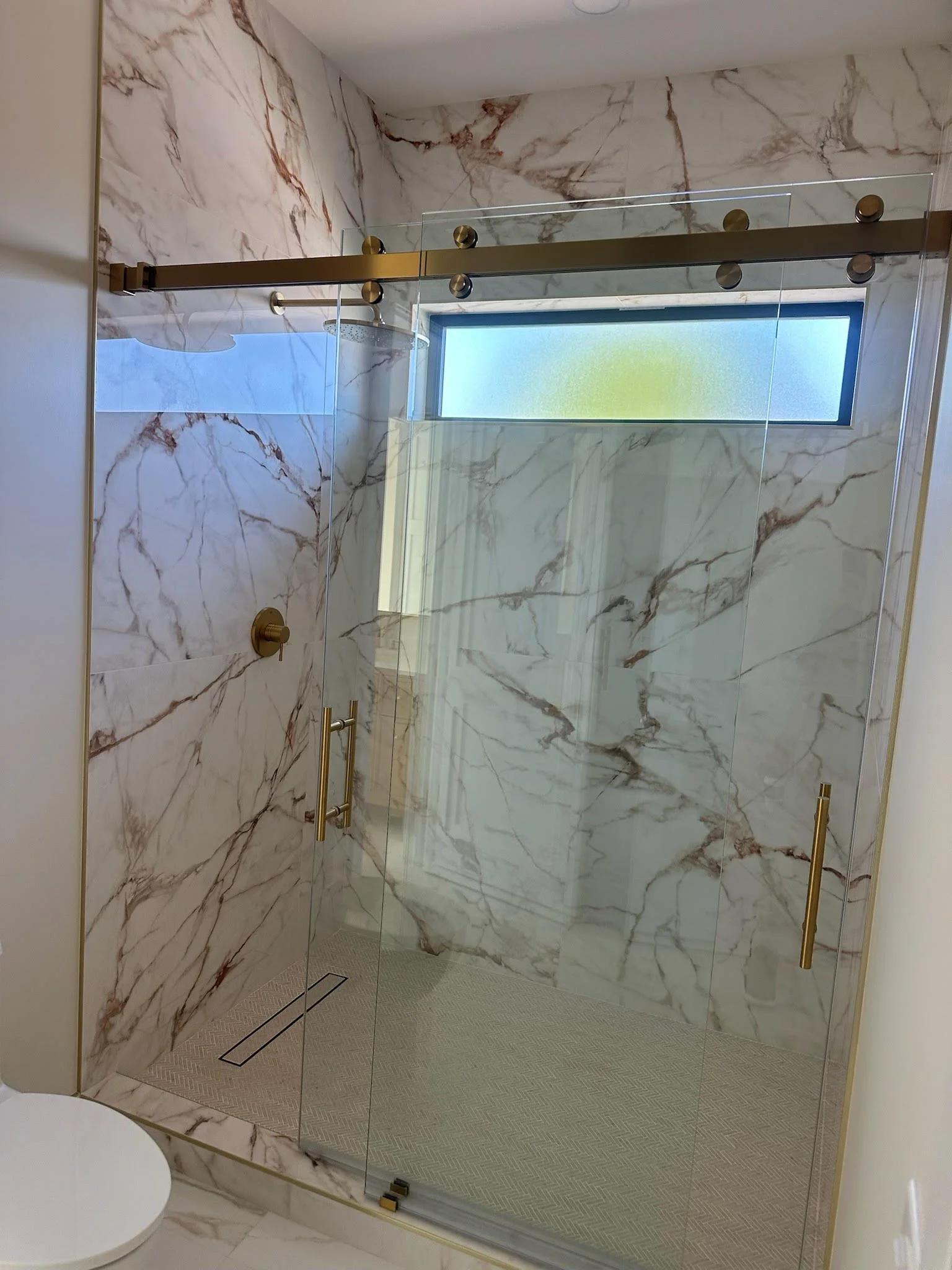 A modern walk-in shower with glass doors, gold fixtures, and marble walls, featuring a horizontal frosted window.