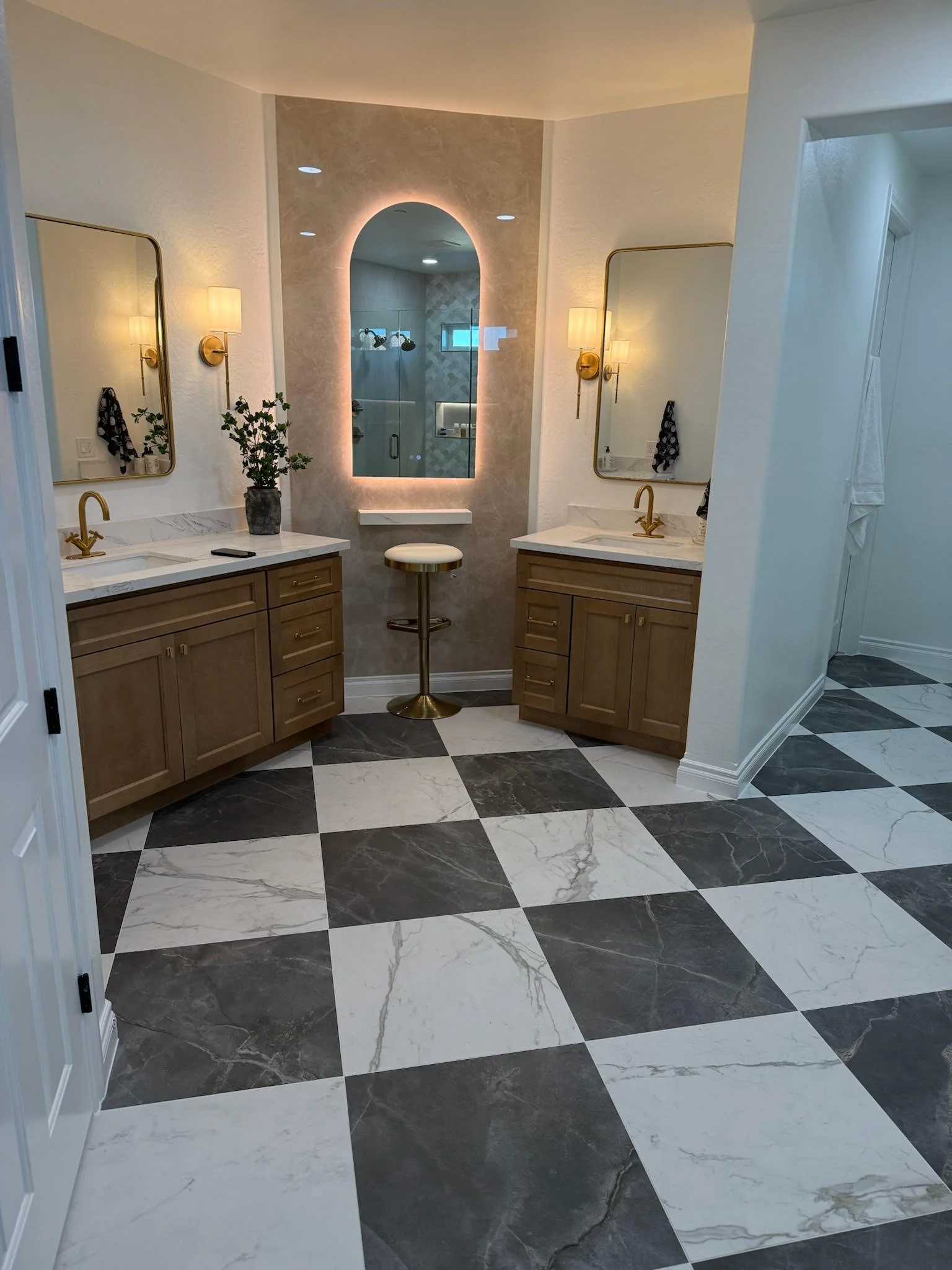 A modern bathroom with two wooden vanities, each with a marble countertop and a gold faucet. Above the vanities are mirrors with gold frames and wall-mounted light fixtures. A plant is on the left vanity, and a black and white towel hangs on a hook. In the center, there is a niche with a backlit mirror and a small stool beneath it. The bathroom features black and white checkered marble flooring.