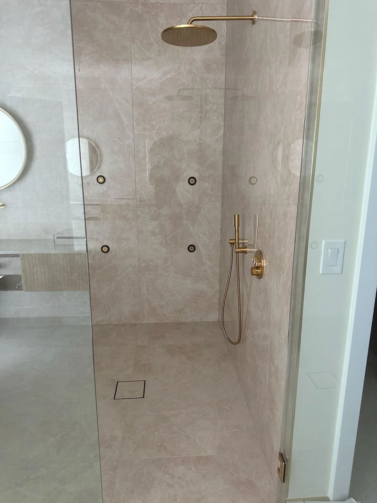 A modern shower with beige marble tiles, a large round gold showerhead, and gold fixtures, including a hand-held shower wand and control valve.