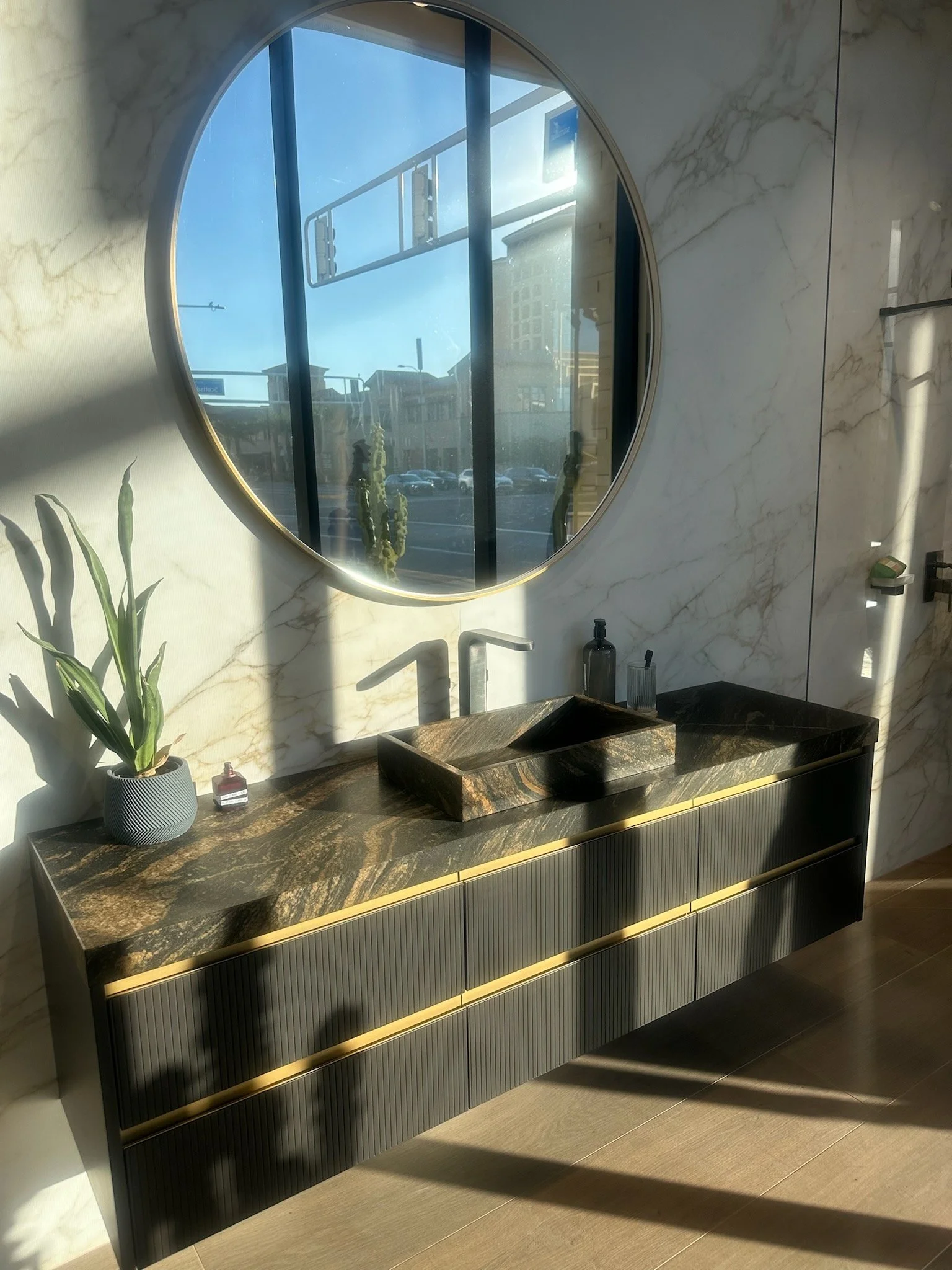 Modern bathroom vanity with a marble countertop, a black vessel sink, and a round mirror above it. There are decorative bottles, a potted plant, and a window reflected in the mirror. The wall and floor have marble and tile finishes.