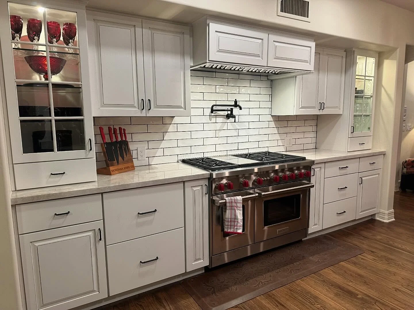 White kitchen cabinets with black handles, white tiled backsplash, stainless steel stove with red control knobs, red and white checkered towel, knife set on countertop, glass-front cabinets with red glassware inside, and hardwood flooring.
