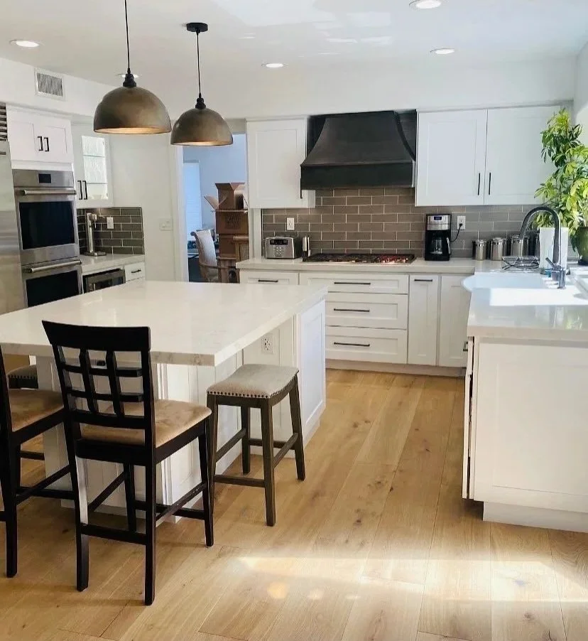 Modern kitchen with white cabinets, a large white island, black bar stools, stainless steel appliances, black range hood, and wood flooring.