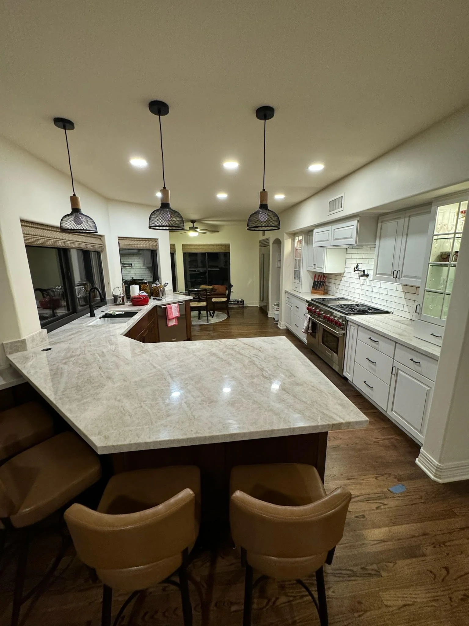 Modern kitchen with white cabinets, marble countertop, stainless steel appliances, and pendant lighting. There's a breakfast bar with brown chairs and a dining area with a table and chairs in the background.