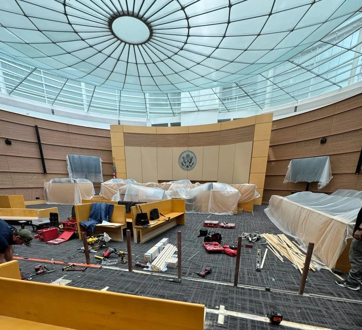 The interior of a large, modern government building or courtroom under construction, with wooden paneling, a circular glass ceiling, and a symbol of the United States on the wall, surrounded by construction tools and covered furniture.