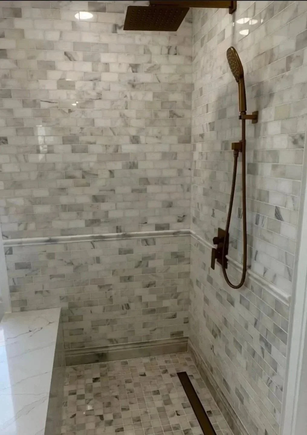 A walk-in shower with white marble tile walls, a walk-in bench, and bronze fixtures including a rainfall showerhead and a handheld shower wand.