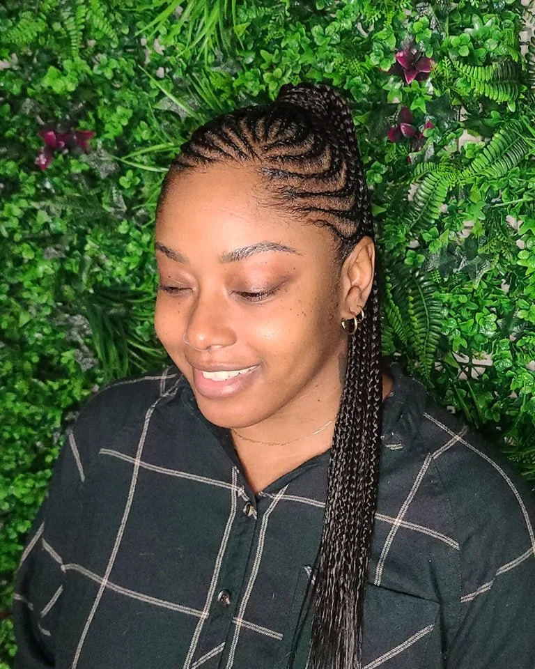 A woman with braided hair, wearing a black shirt with a checkered pattern, standing in front of a lush green, leafy background.