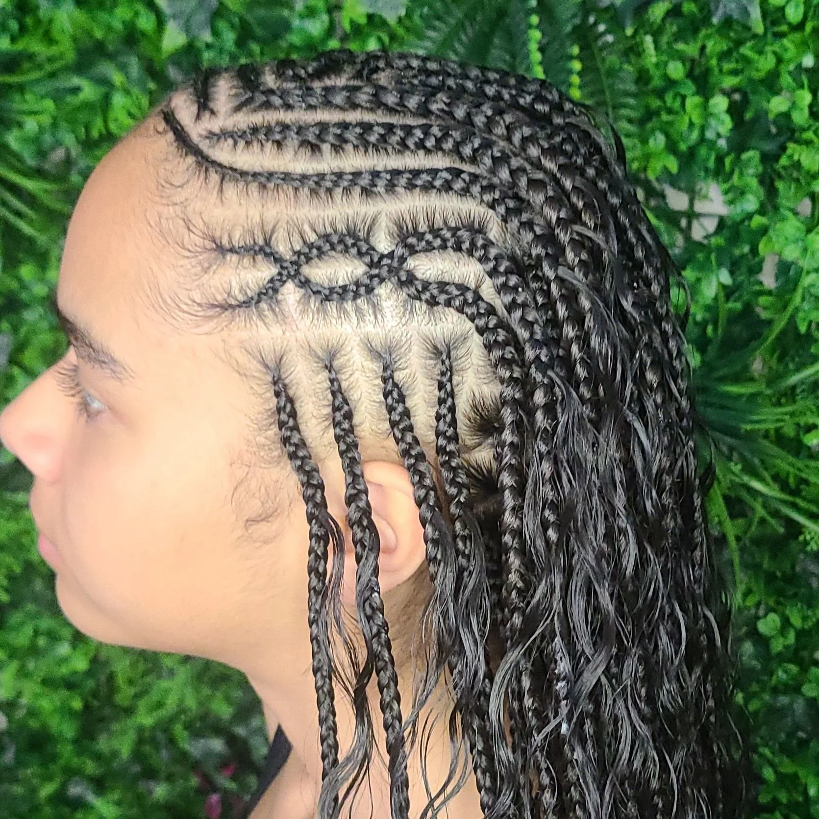 Side view of a woman with intricately braided hair, featuring various patterned cornrow styles, in front of green foliage.