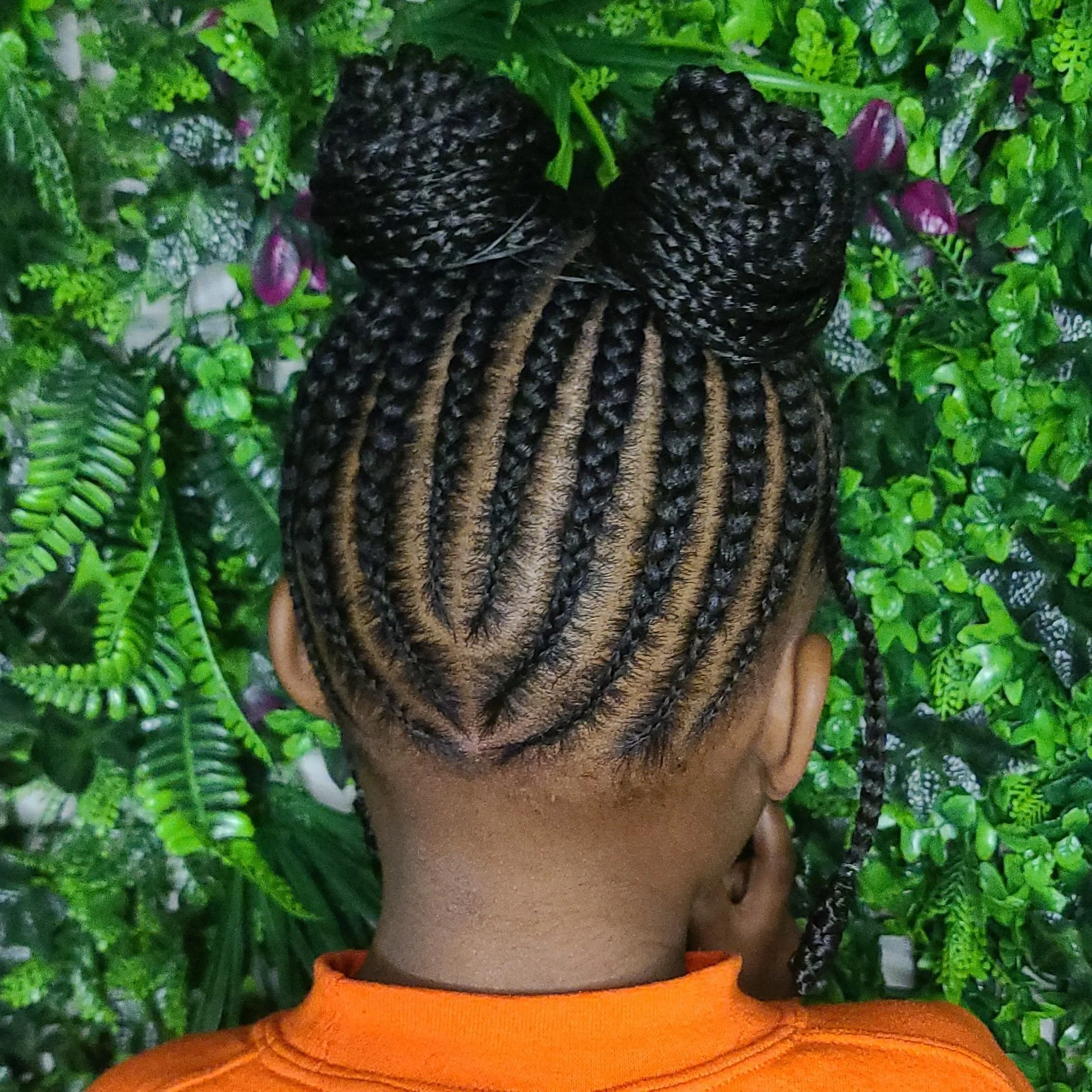 Back view of a person with braided hair styled into two buns and cornrows, wearing an orange shirt, in front of green foliage.