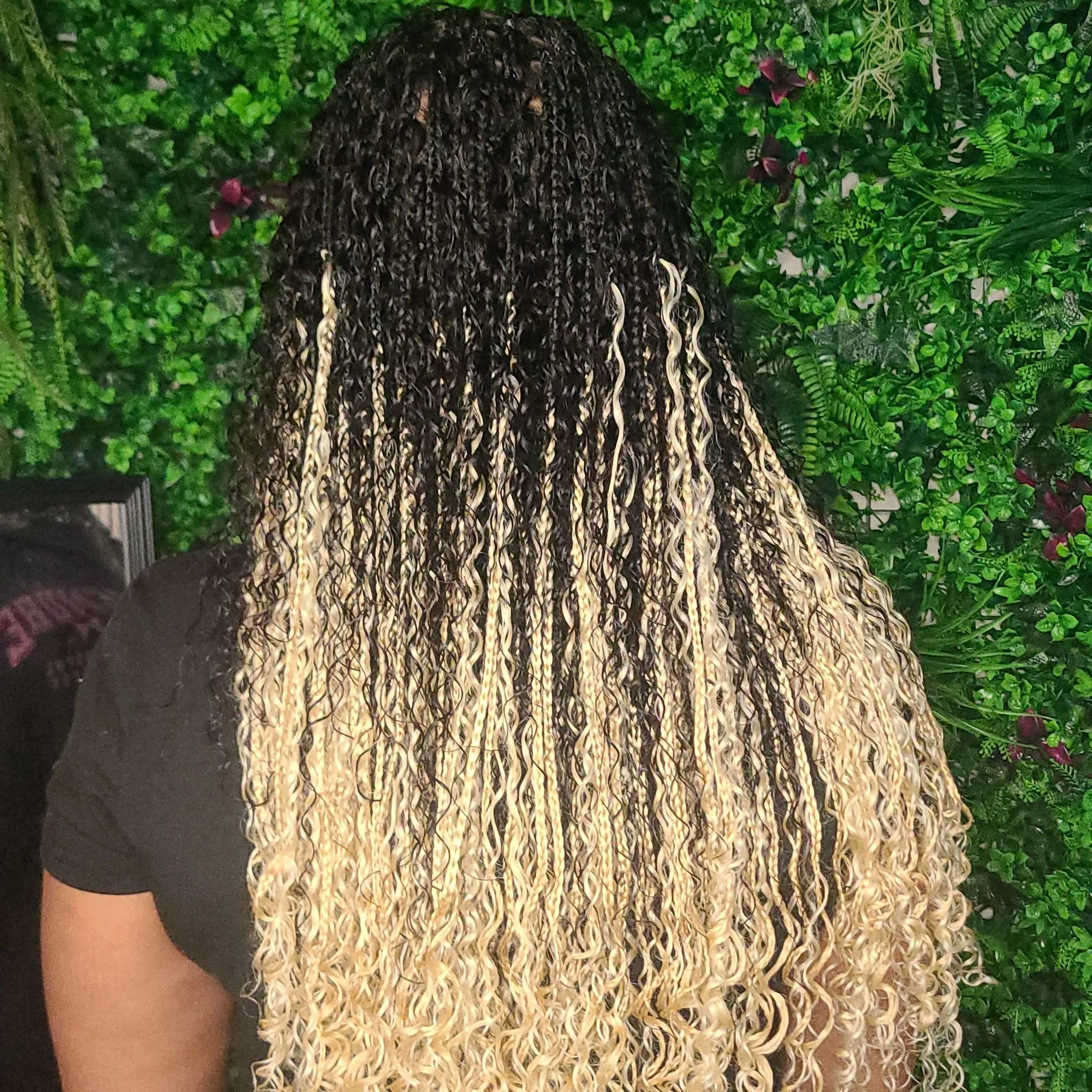 Back view of a woman with long, curly hair with black roots and blonde tips, standing against a green leafy background.