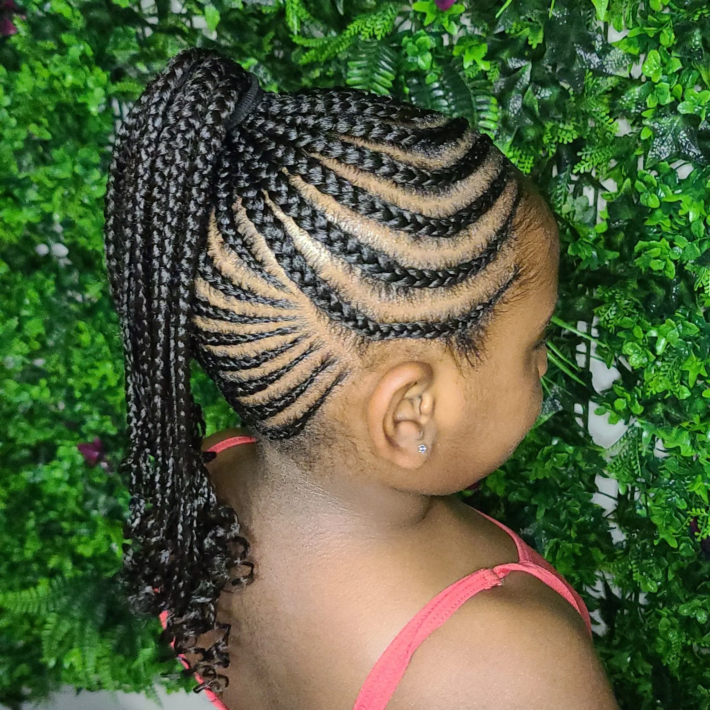 A woman with intricately braided hairstyles featuring thin, long braids styled in a high ponytail with curly ends, standing against a green leafy background.