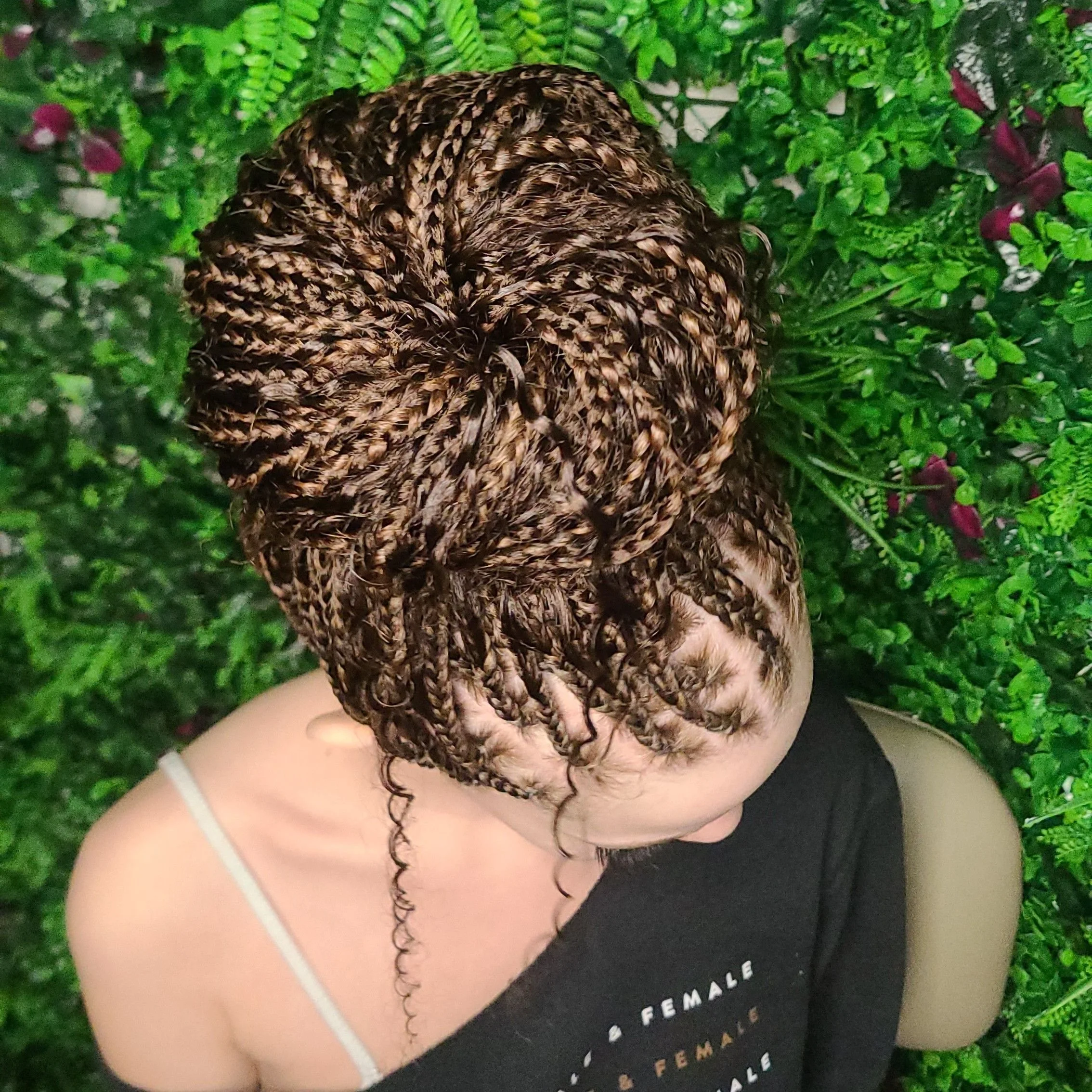 A woman with intricately styled small, twisted braids, some forming a bun, surrounded by lush green foliage.