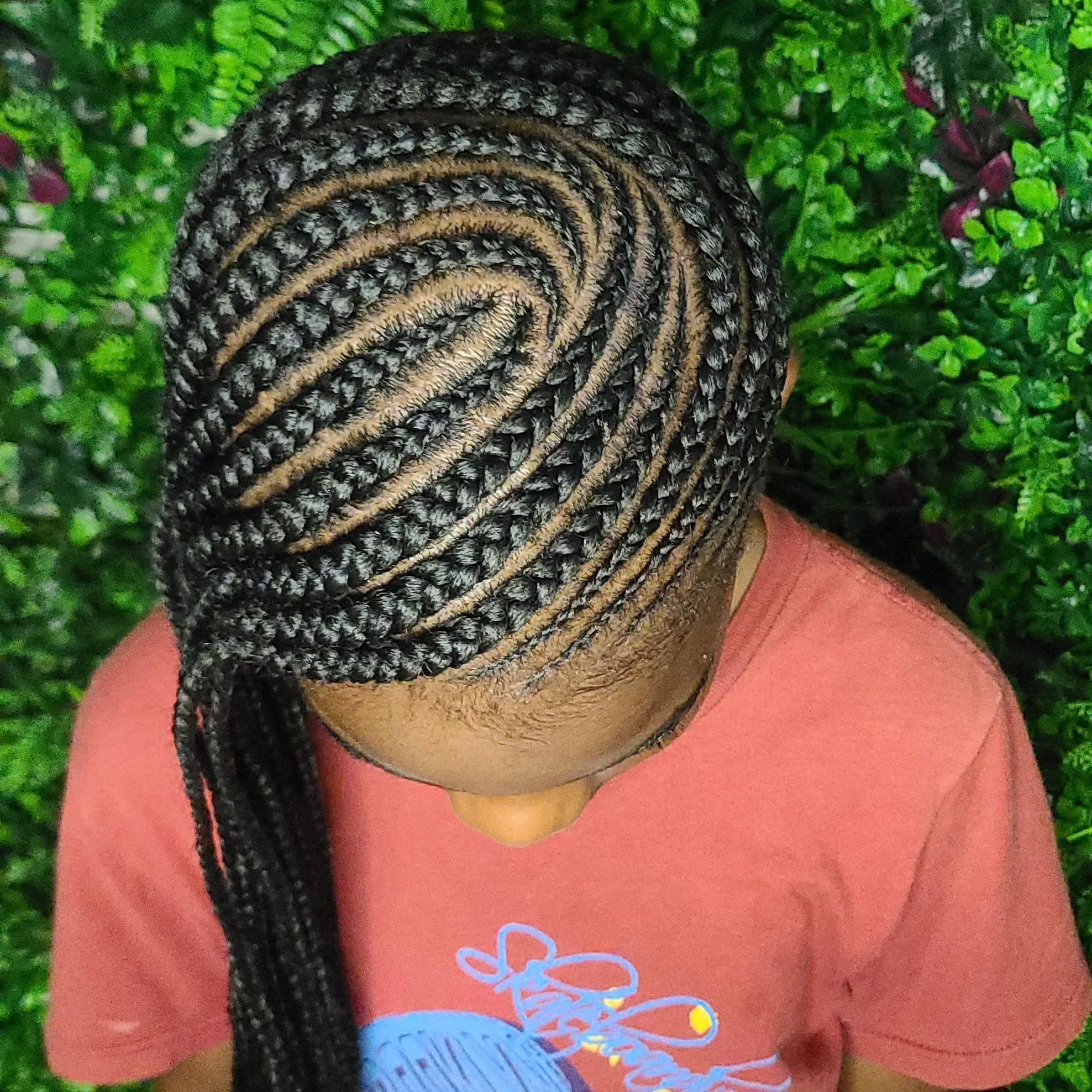 Top view of a person with intricately styled braided hair, wearing a pink shirt, and standing in front of green plants.