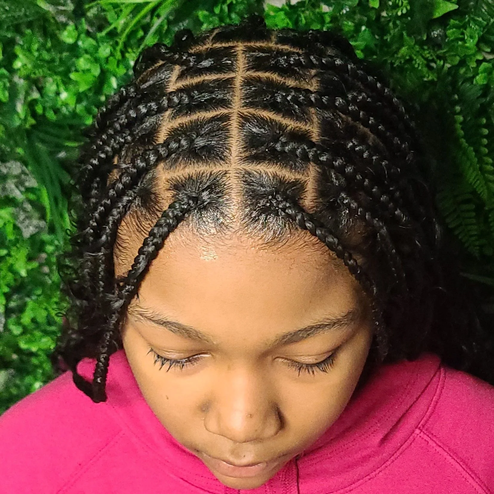 Top view of a woman with neatly styled box braids on her head, lying on green foliage, wearing a pink top.