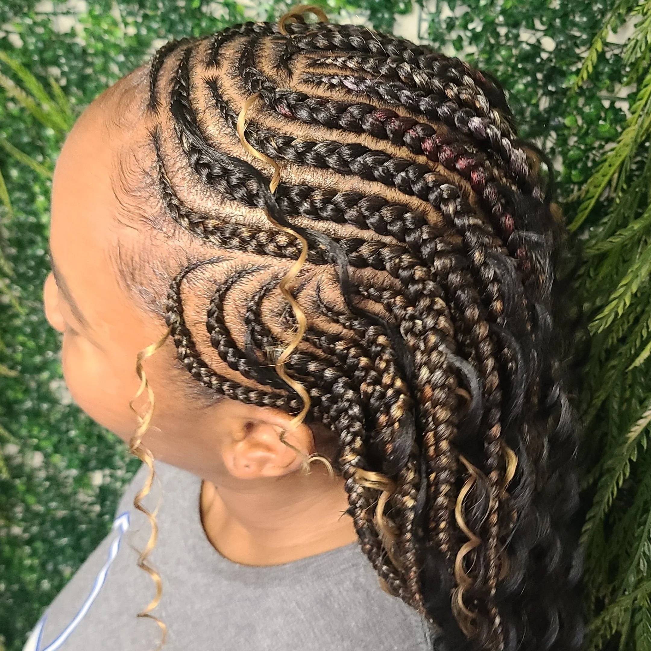 Close-up of a woman with a new intricate braided hairstyle with blonde curly accents, set against a leafy green background.