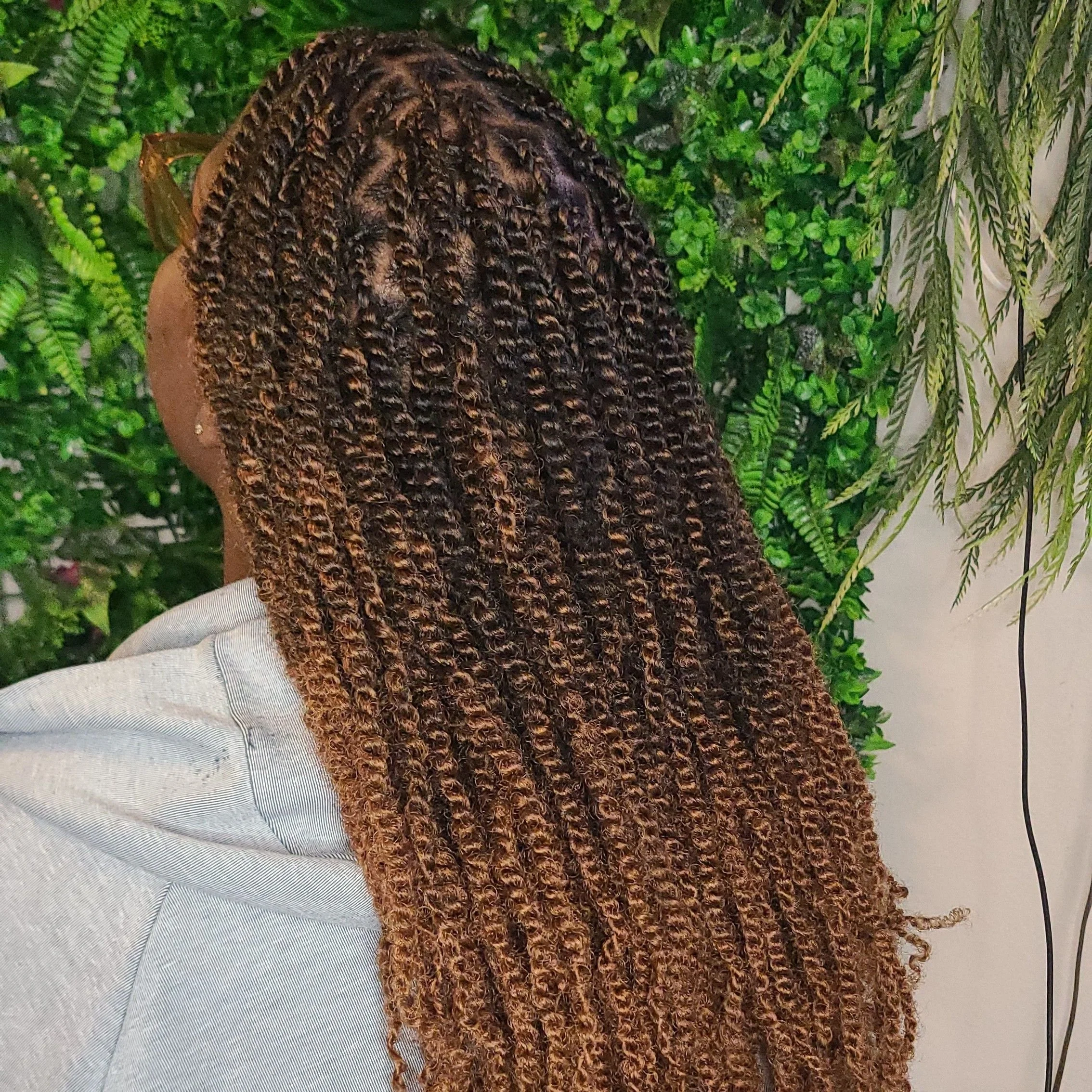 Person with long, styled curly hair, wearing glasses and a gray hoodie, standing in front of green foliage.