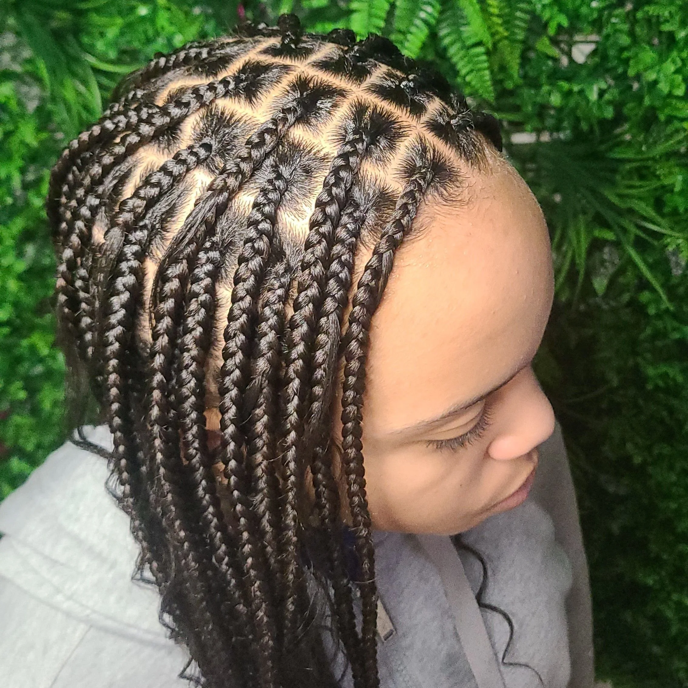 Close-up of a woman with neatly done black box braided hairstyle, surrounded by green foliage.