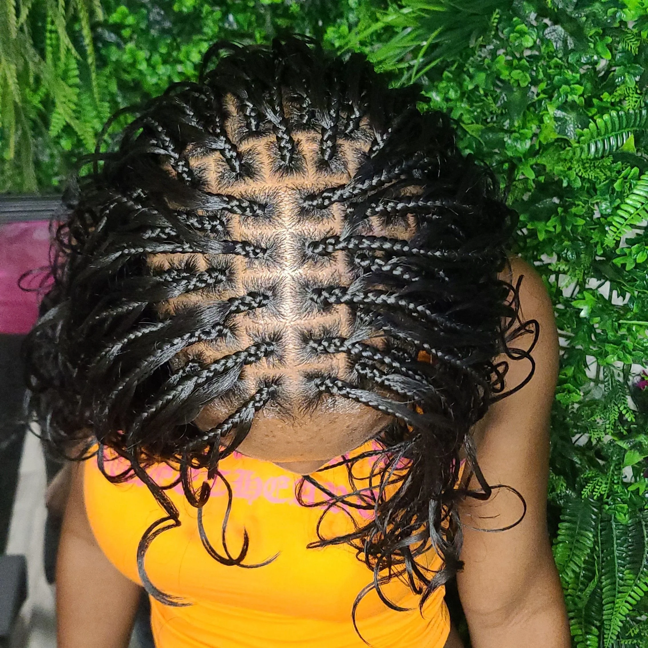 Top view of a person with wet, styled black and white twisted braids, wearing an orange top, standing in front of green plants.