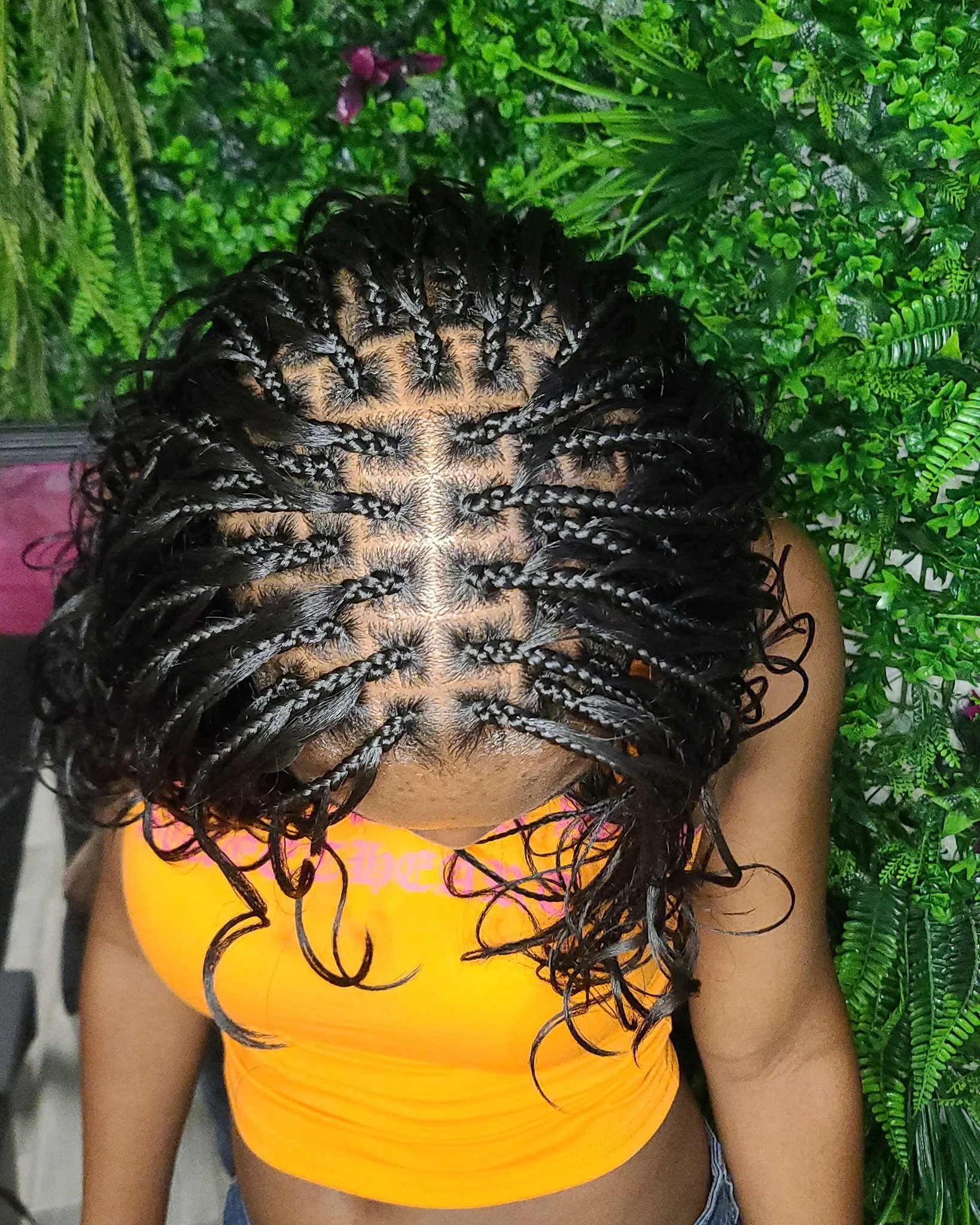 Top view of a woman with curly, braided hair, wearing an orange shirt, standing in front of green foliage.