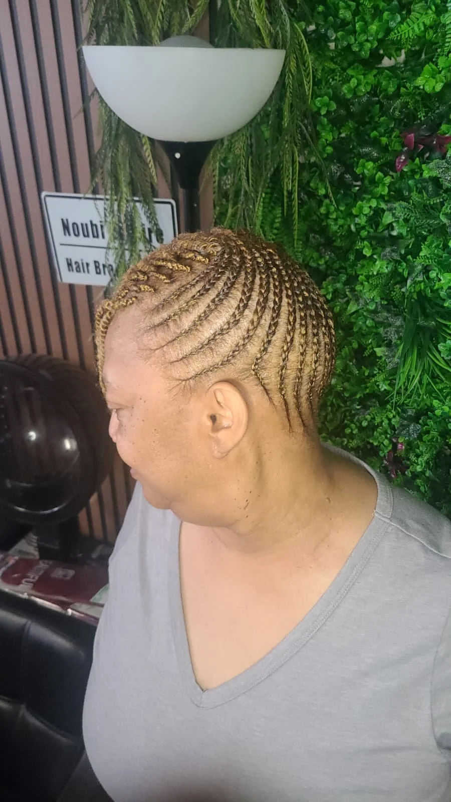 A woman with blonde cornrow braids in a salon with green plants in the background.