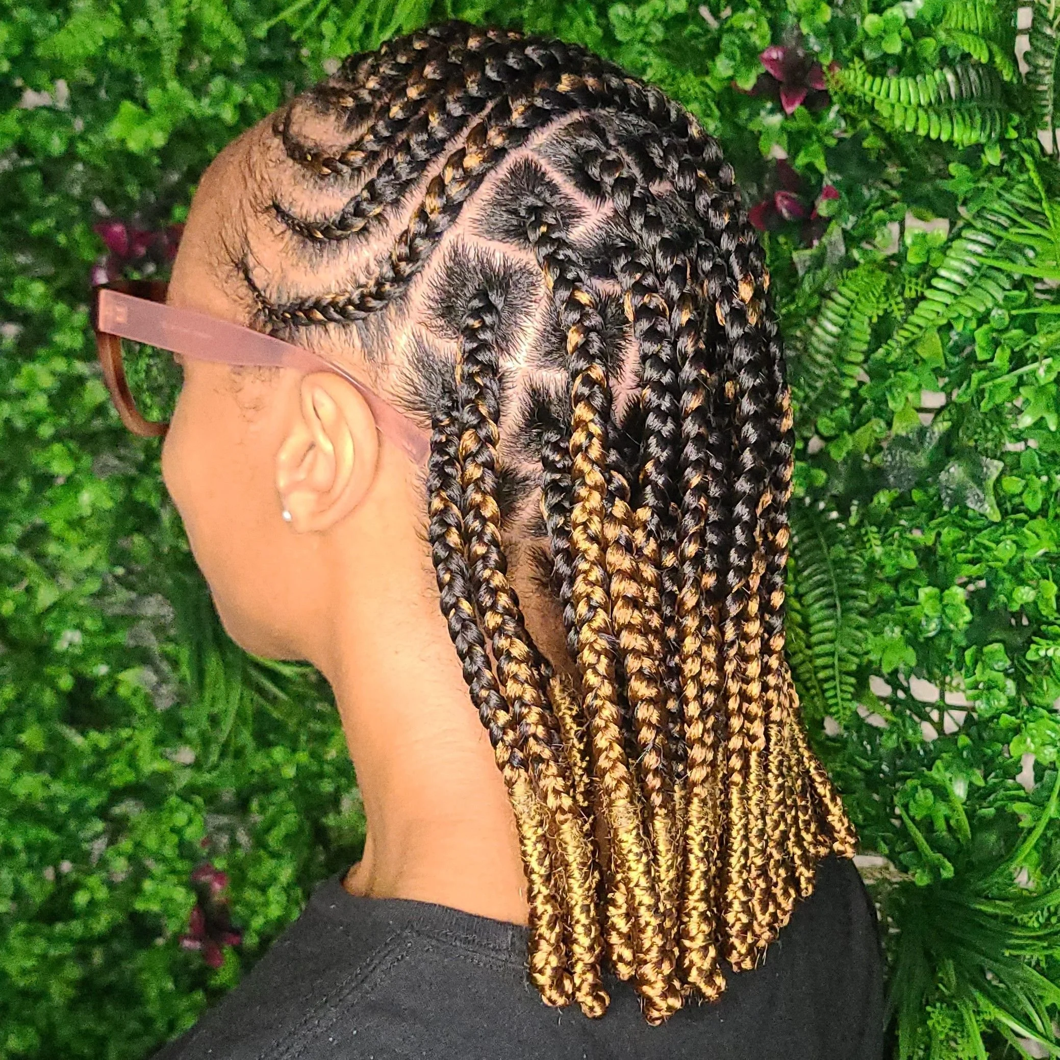 Side view of a woman with intricately braided hairstyle featuring black and blonde braids, wearing glasses, against a backdrop of lush green plants.