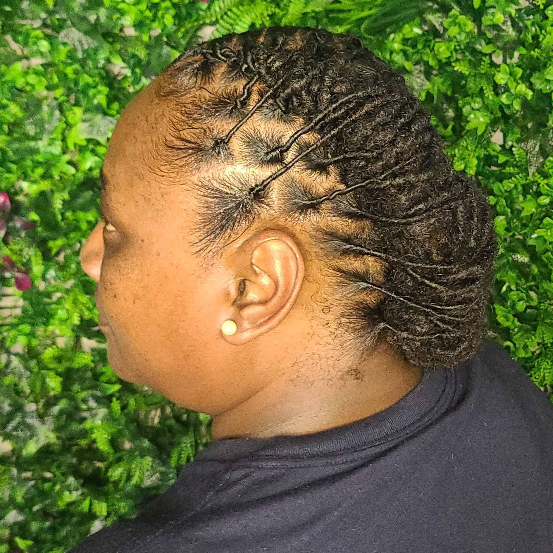 A woman with intricately styled hair in braids and twists, wearing pearl earrings, against a green leafy background.