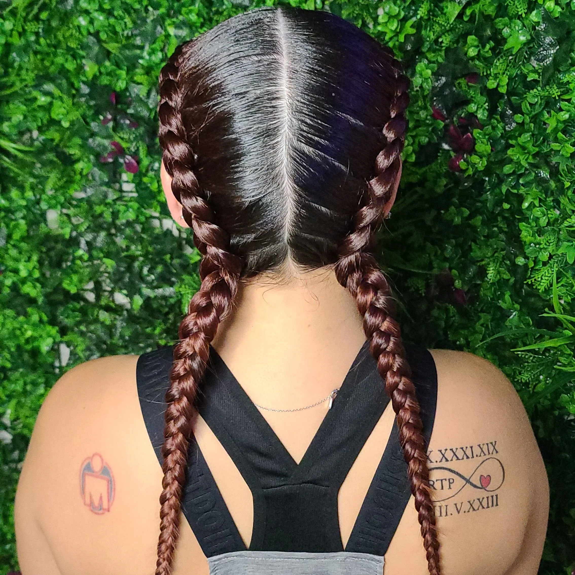 A person with long, dark hair styled in two braids, wearing a black top with cut-out shoulders, standing against a background of green foliage. They have tattoos on both shoulders; the left tattoo is a small, stylized person, and the right tattoo fea