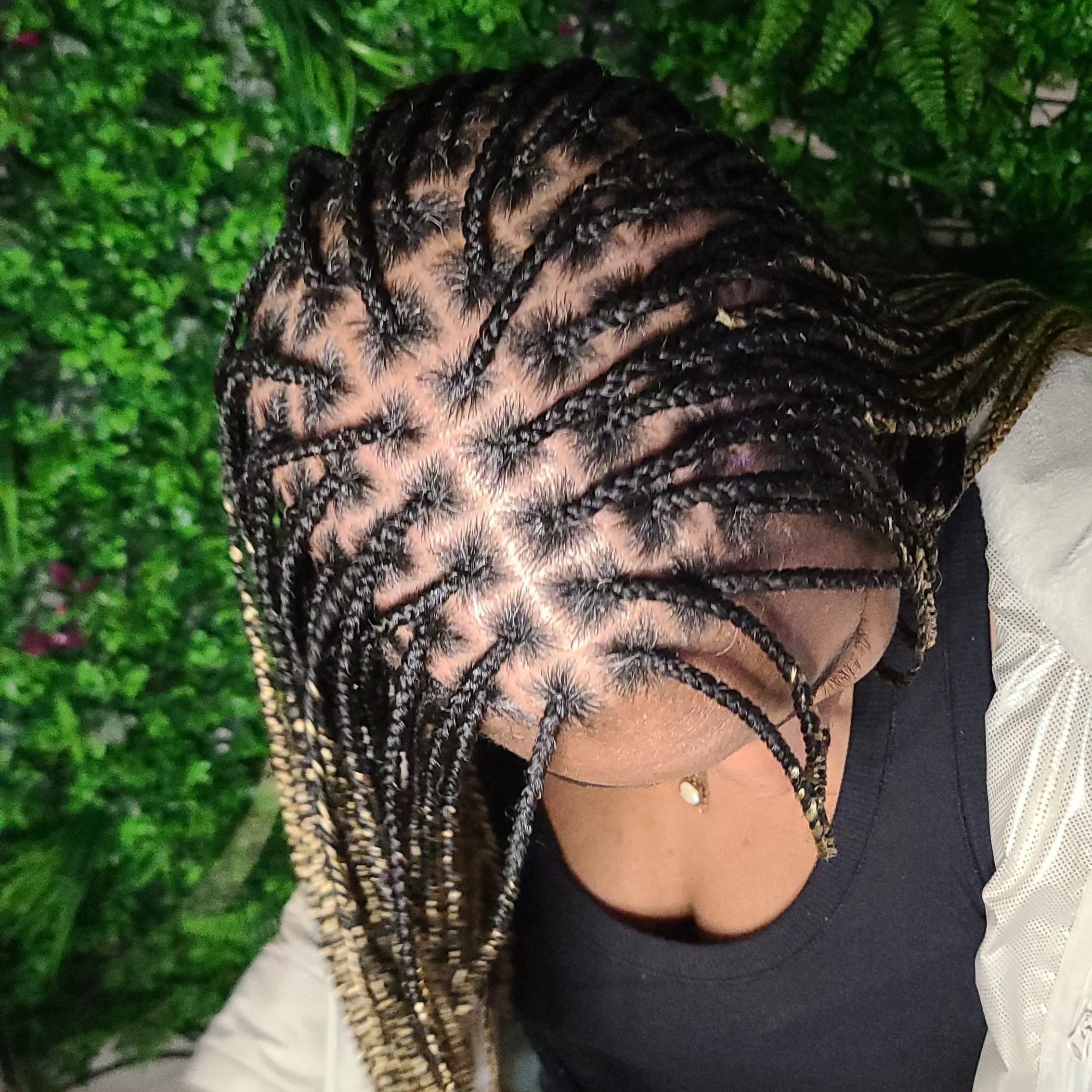 A woman with long, braided hair, mostly in black with light blonde accents, looking down. She is wearing a black top and a light-colored jacket, with greenery in the background.