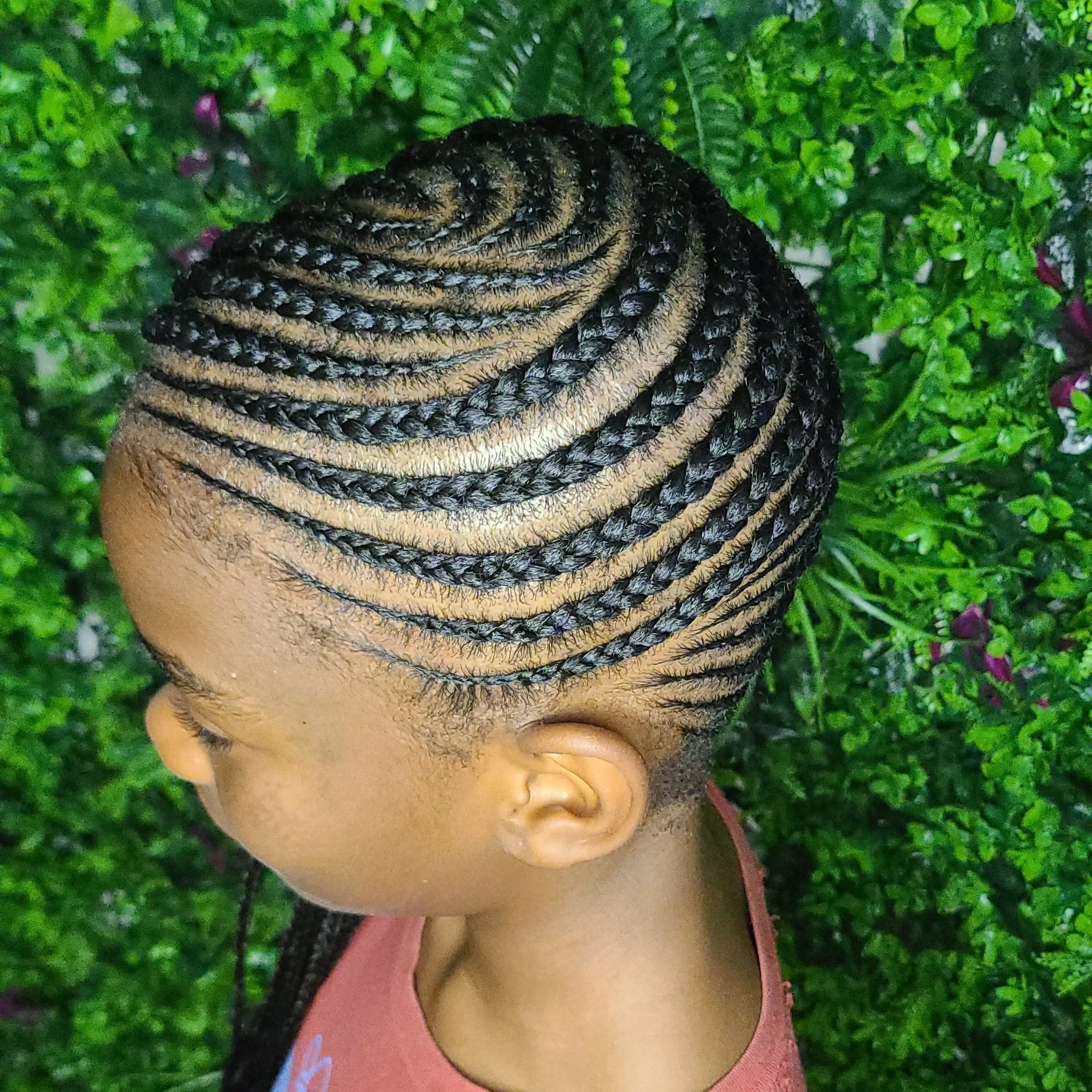 A side view of a person with neatly braided hair styled in cornrows, with alternating black and gold-colored braids, against a background of green foliage.