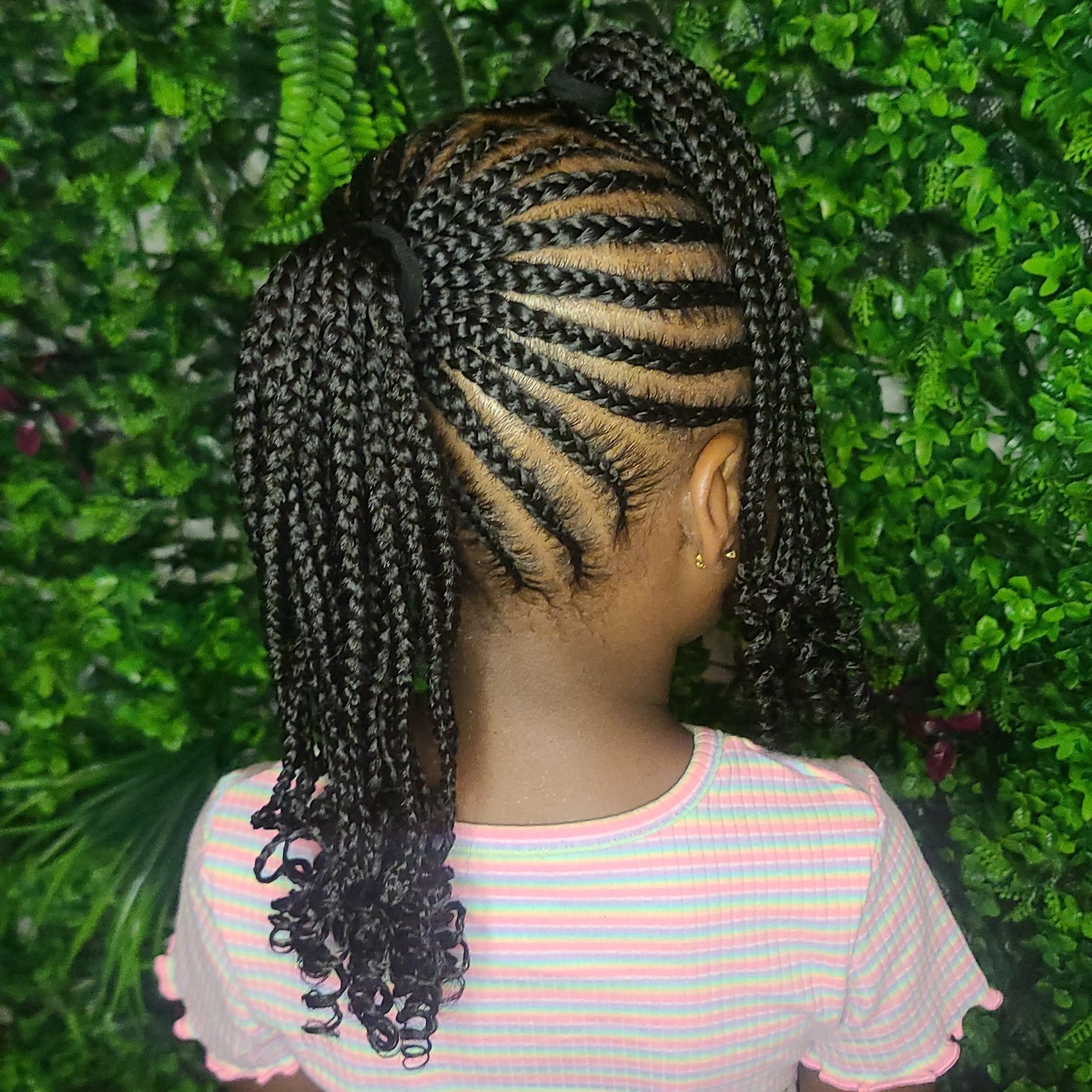 A woman with intricately braided hair styled in a partial updo with curls at the ends, standing in front of lush green foliage, wearing a pink and white striped shirt.