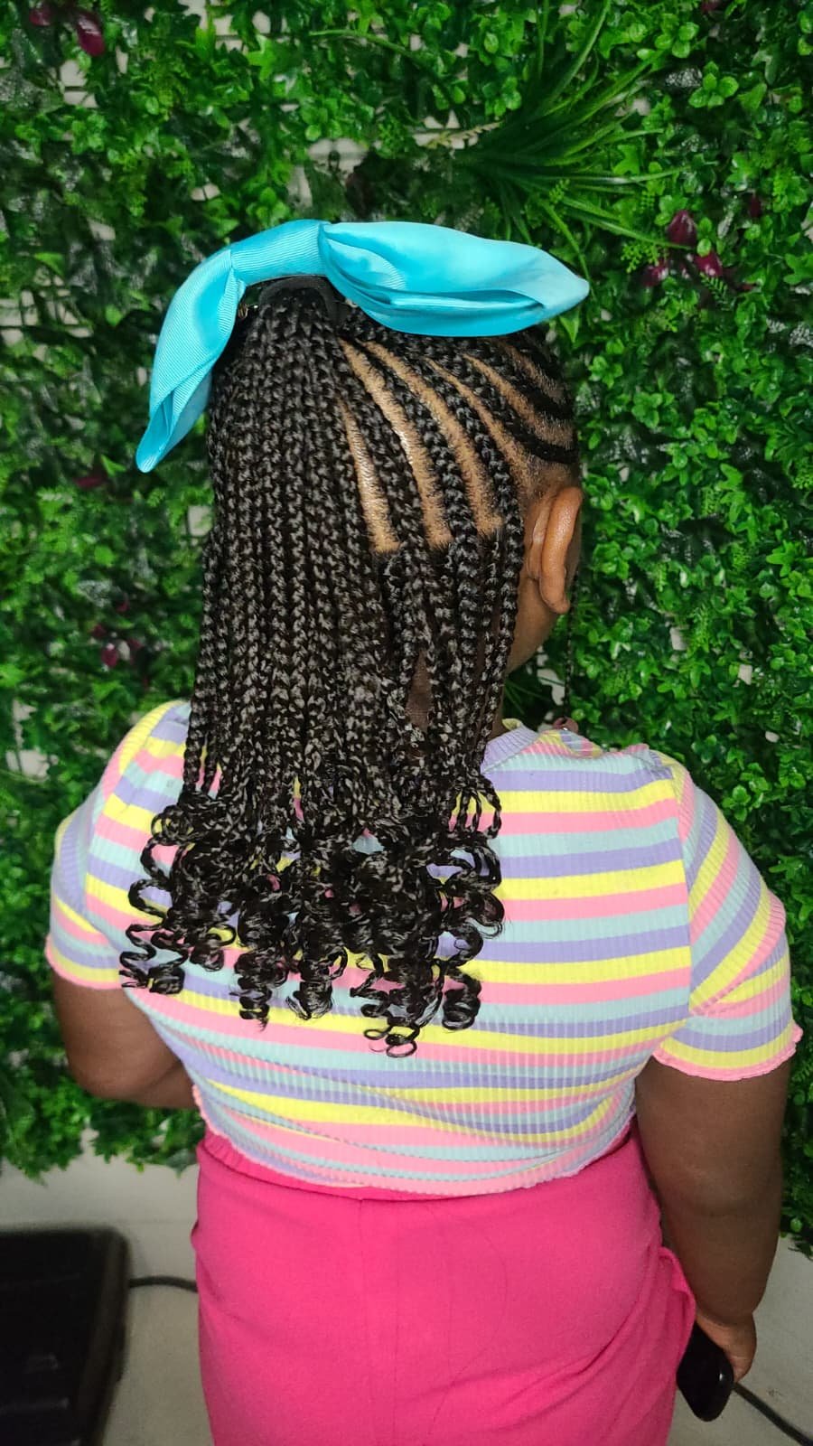 Back view of a woman with braids and curls wearing a striped pastel shirt and pink skirt, standing in front of a green leafy wall with a blue headband.
