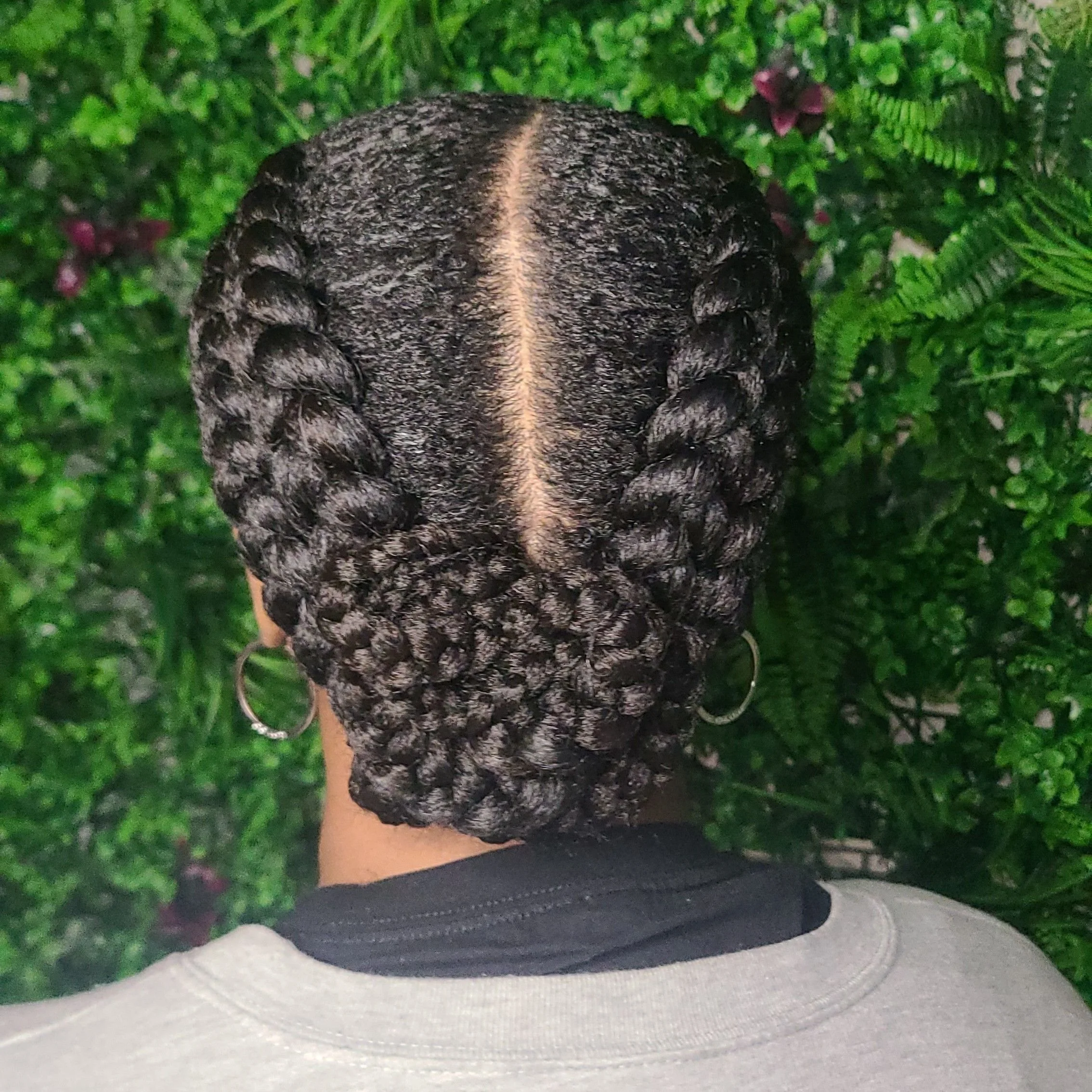 Back view of a person with neatly styled box braids arranged in a crown-like updo, with a middle part, against a background of green plants and ferns.