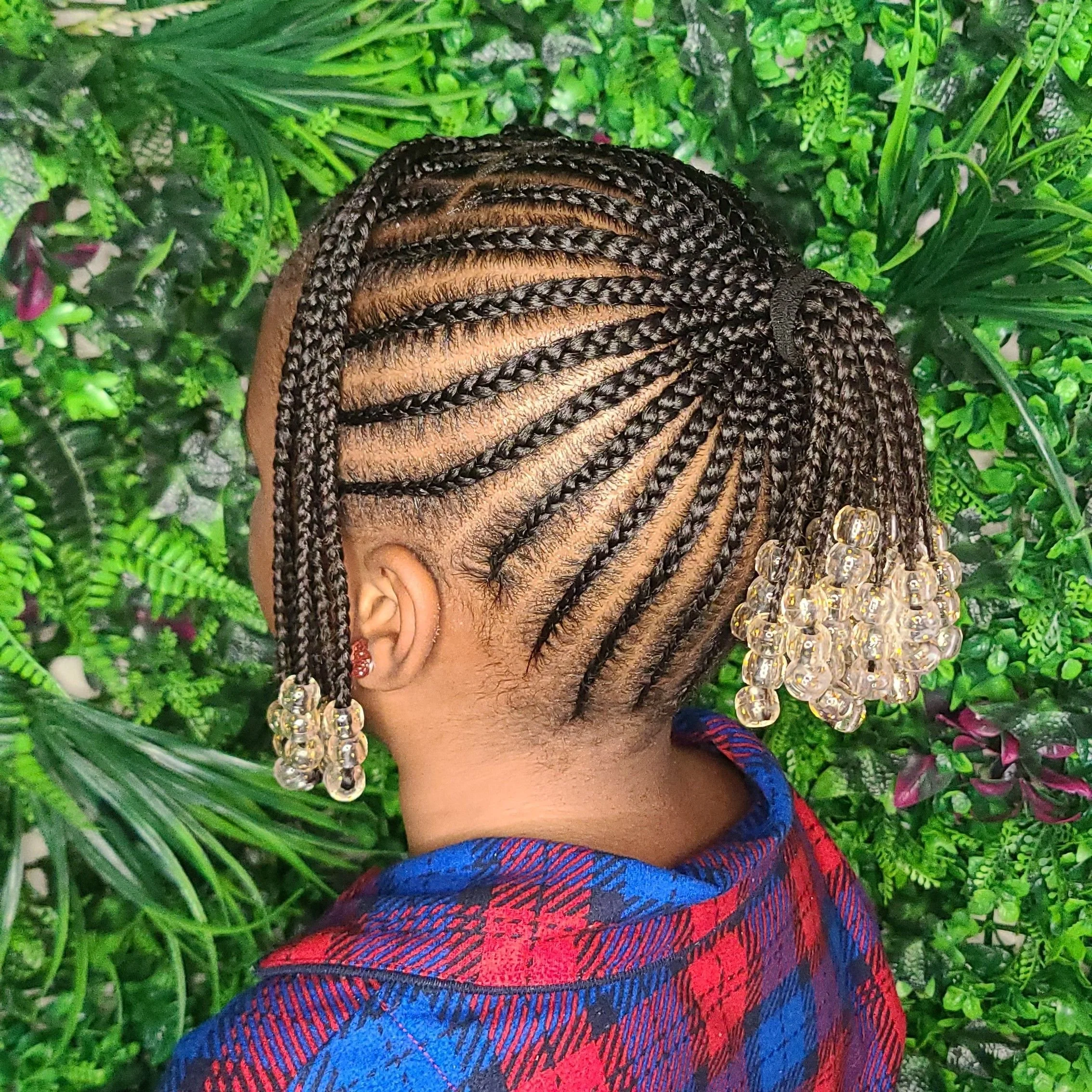 Close-up of a woman with braided hairstyle decorated with clear bead accessories, wearing a red and blue plaid shirt, standing against a backdrop of lush green foliage.