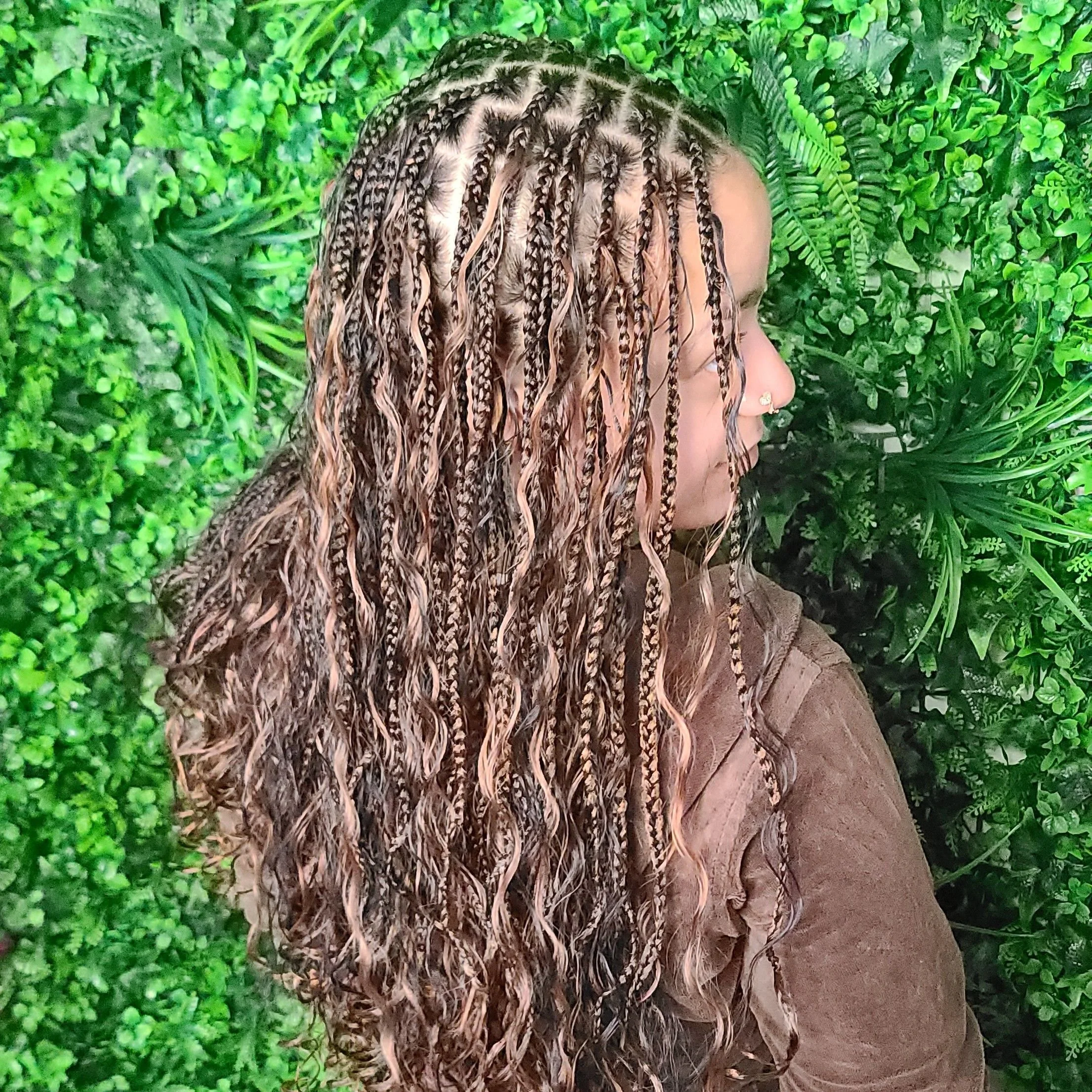 Side profile of a woman with long, curly, and braids hair standing in front of a lush green leafy background.