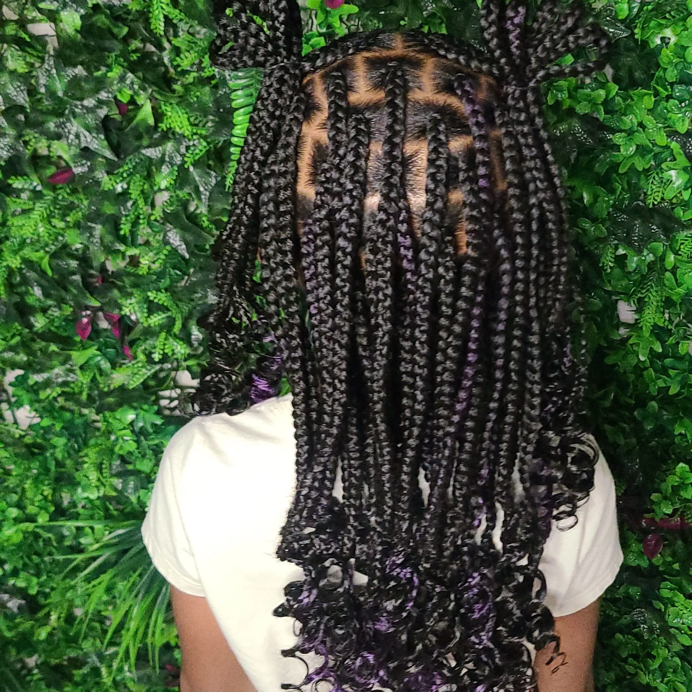 Person with long, curly, twist-style braids standing against a lush green leafy background.