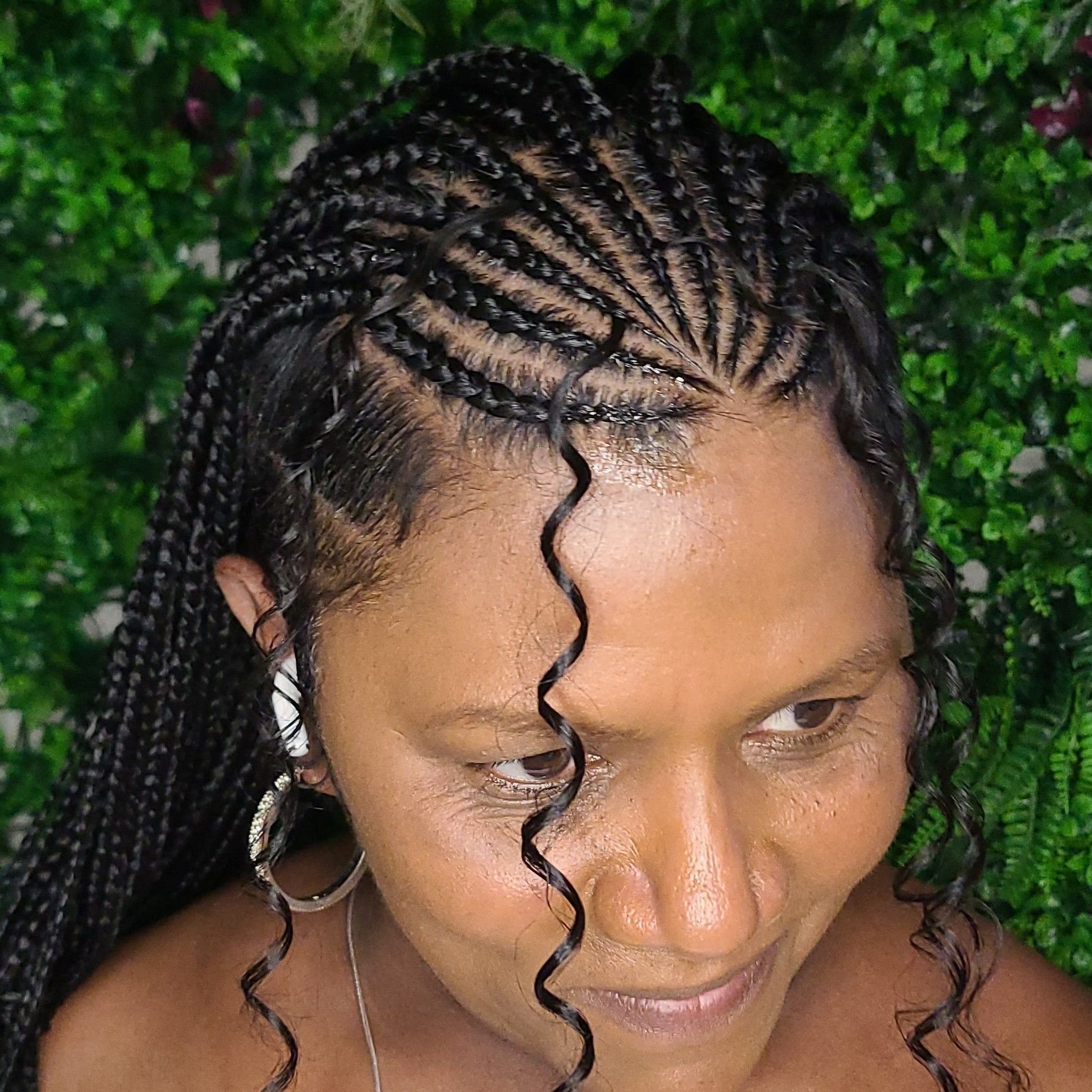 A woman with braided hairstyle that includes one loose curly strand in the center, wearing hoop earrings, smiling softly, with green leafy background.