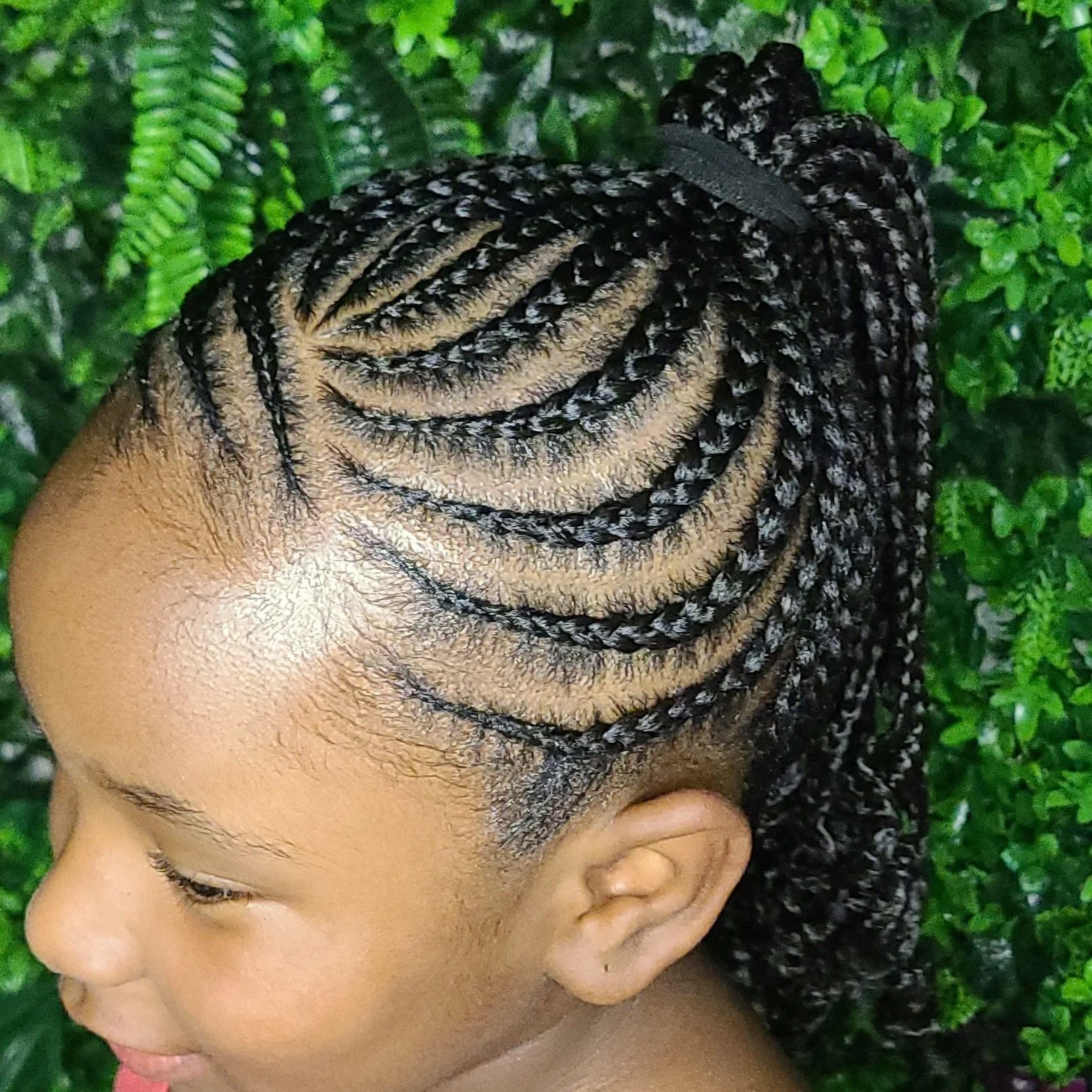 A young girl with braided hair styled in a side-swept updo with black and blonde braids, standing outdoors in front of green foliage.