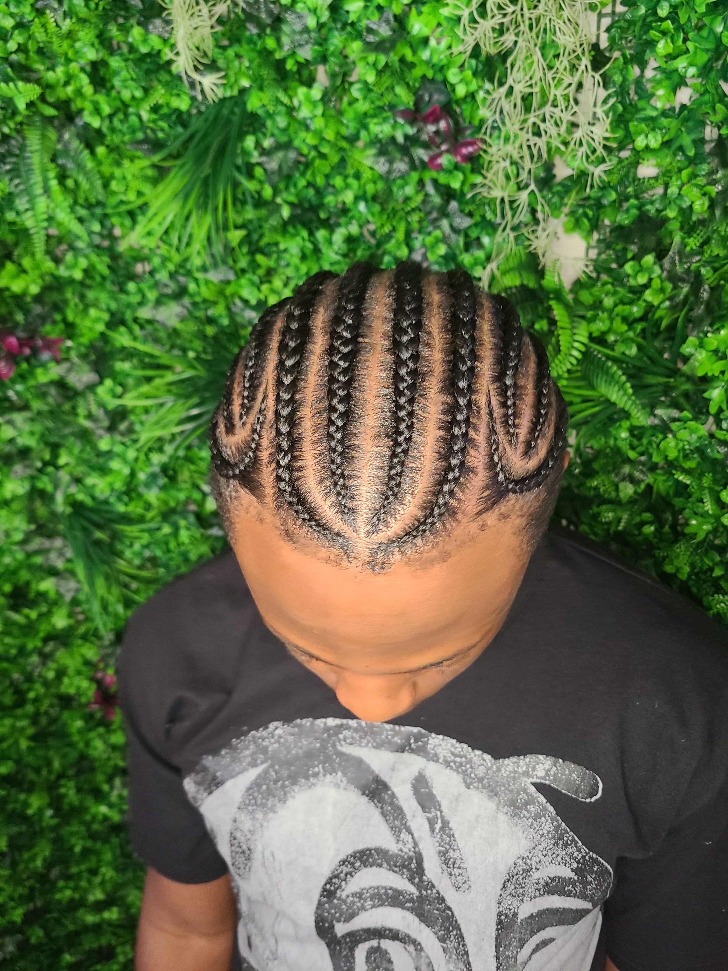 Top-down view of a person with intricately braided hairstyle featuring multiple cornrows, set against a lush green leafy background.