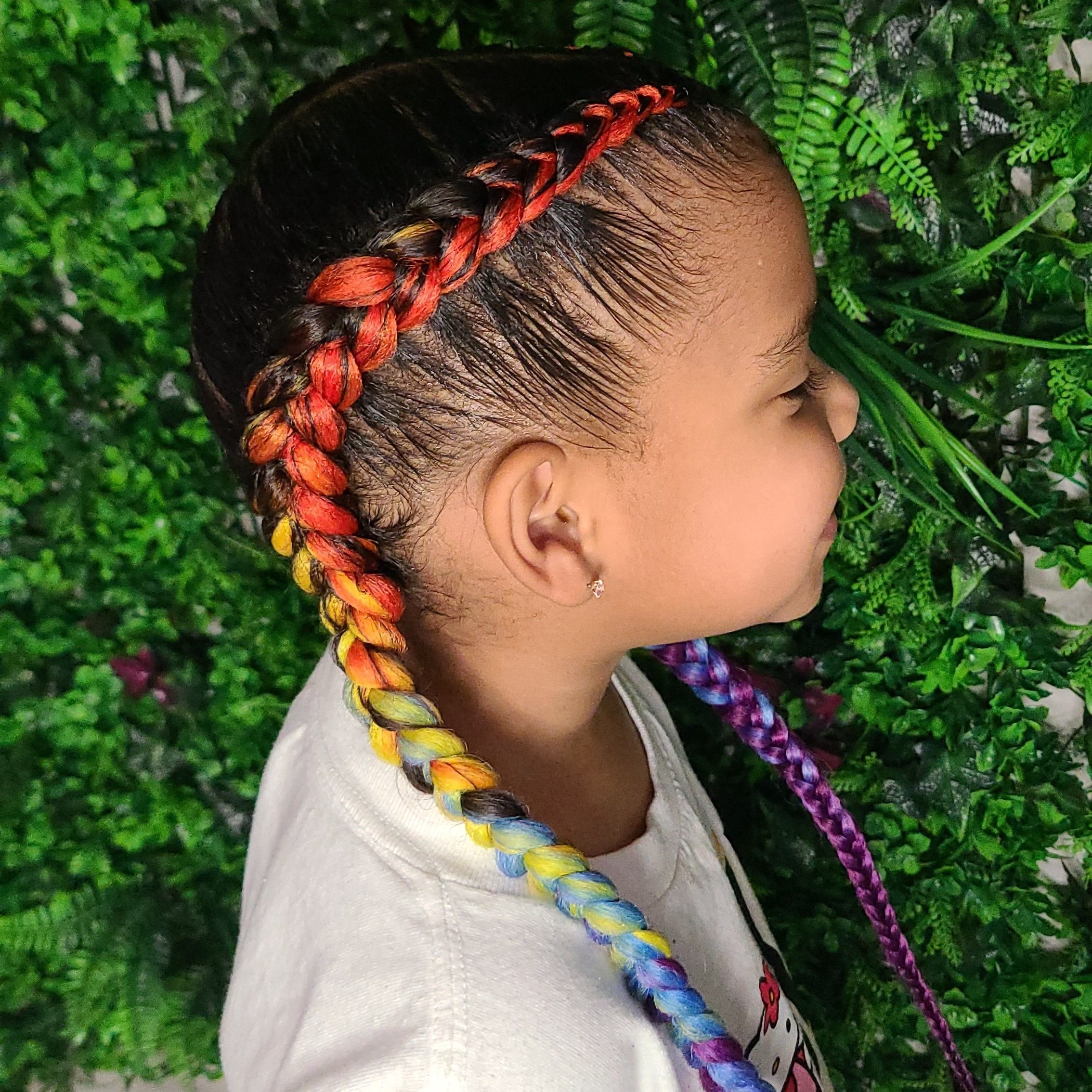 A young girl with a colorful braided hairstyle featuring shades of red, orange, yellow, blue, and purple, standing against a backdrop of green foliage.